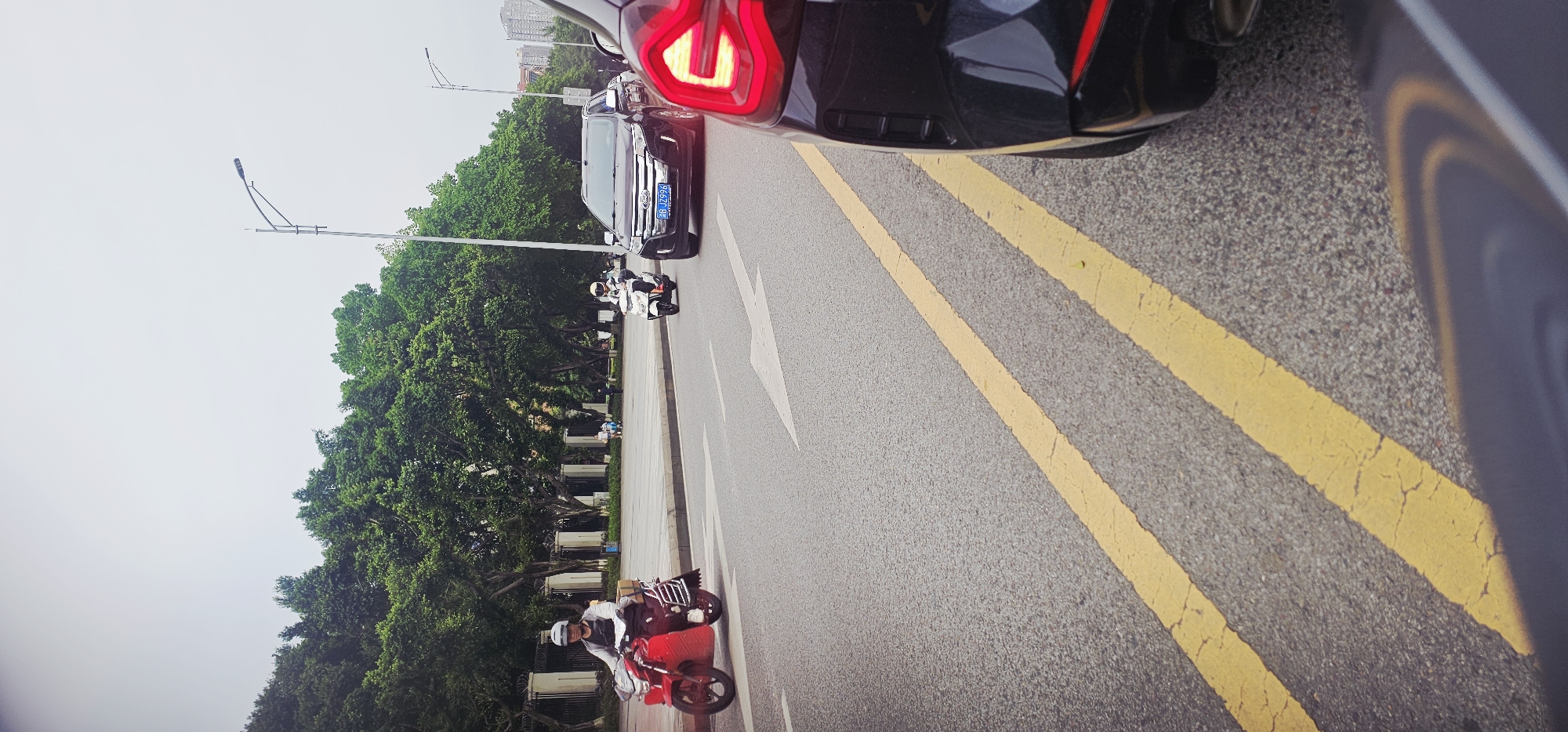 The image shows a street scene in Jiulongpo District, China. A red electric scooter with a rider wearing a white helmet and carrying items on the back is traveling on the road. A dark-colored SUV with a visible license plate is in front of the scooter. The road has yellow double lines and arrow markings. Lush green trees line the side of the road. Street lights are visible along the road. Other vehicles are also present on the road. The sky appears overcast.