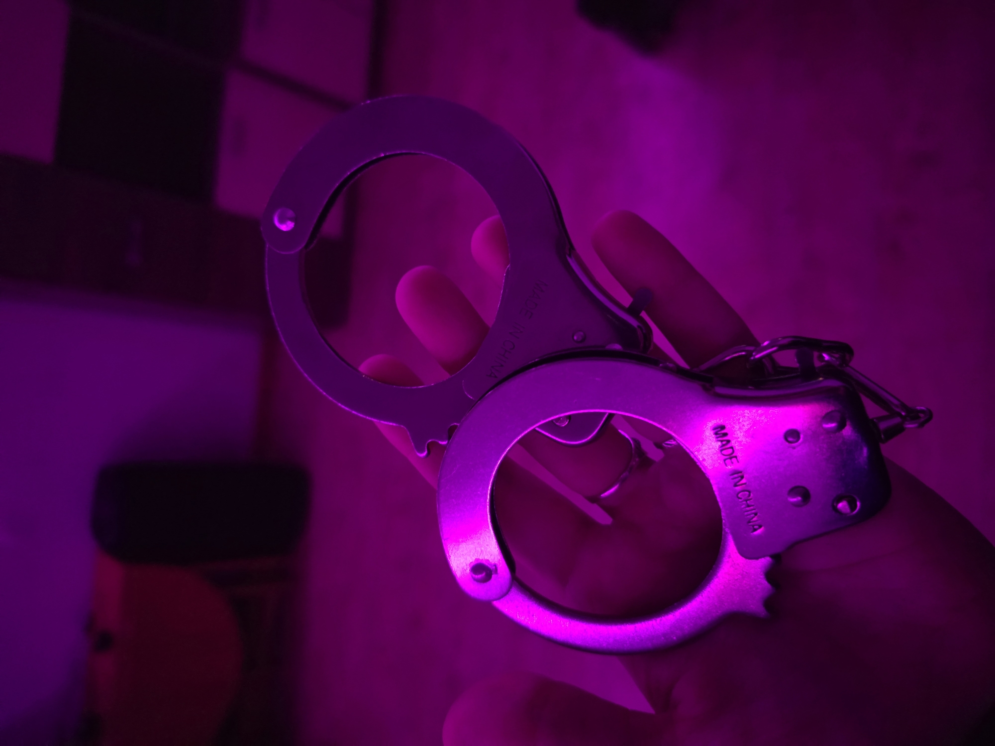 A pair of metal handcuffs is held in a person's hand. The background is blurred, but a shelf-like structure and a cylindrical object are visible. The scene is lit with a purple light. The inscription "MADE IN CHINA" is visible on one of the handcuff components. No information about the location of Chaikovsky, Russia is discernible from the image.