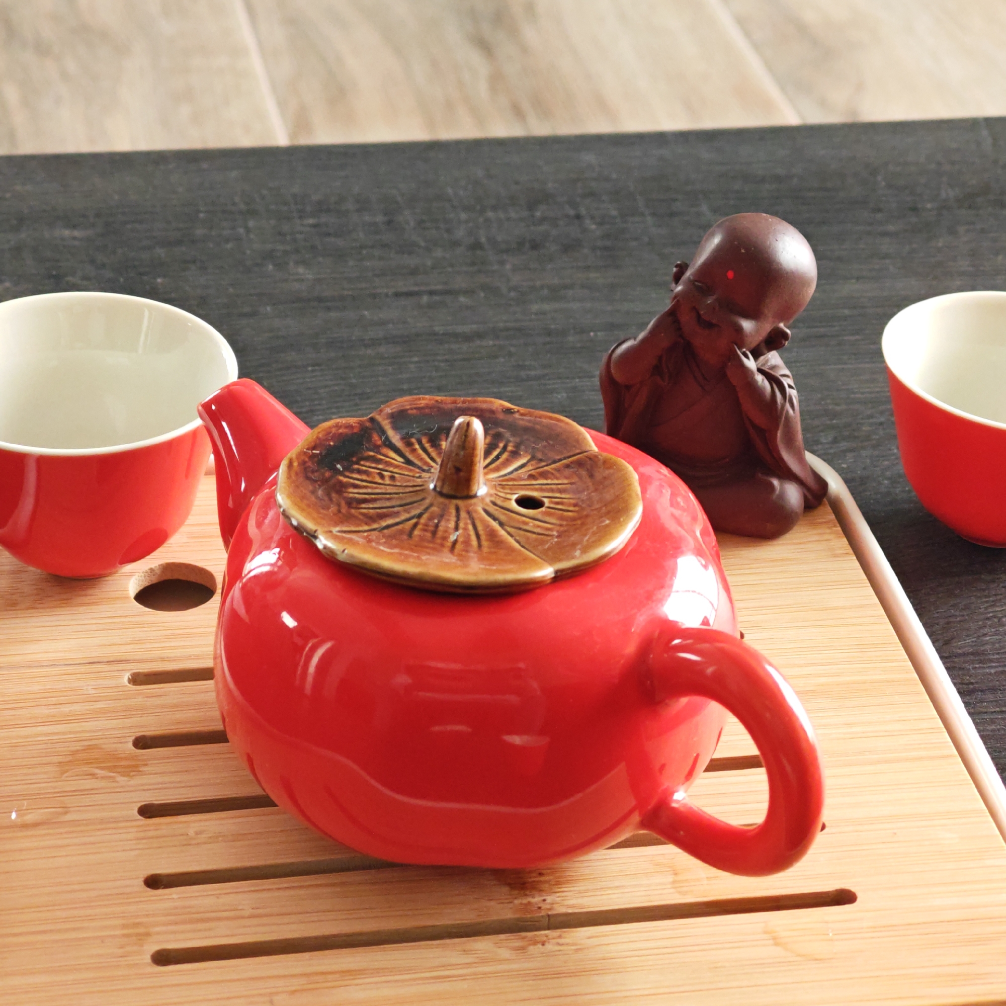A close-up shows a red ceramic teapot with a brown lid placed on a bamboo tea tray, along with two red teacups and a small brown Buddha figurine. The tray is positioned on a dark, textured surface in what appears to be an indoor setting. The objects suggest a tea ceremony or a similar cultural ritual. There is no evidence to suggest the location of this image being specifically in Мысовское сельское поселение, Ukraine.