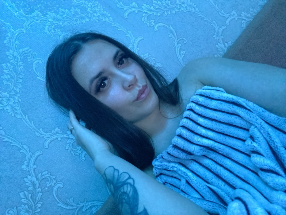 A woman with dark hair and fair skin is lying down, partially covered by a striped, blue towel. She's gazing at the camera with a slight pout. Her left hand rests near her head, sporting a ring. A tattoo is visible on her left arm. The background features a textured, blue wall with a floral pattern. The overall lighting is a cool blue tone, casting shadows and highlighting the details of the towel and the woman's features.