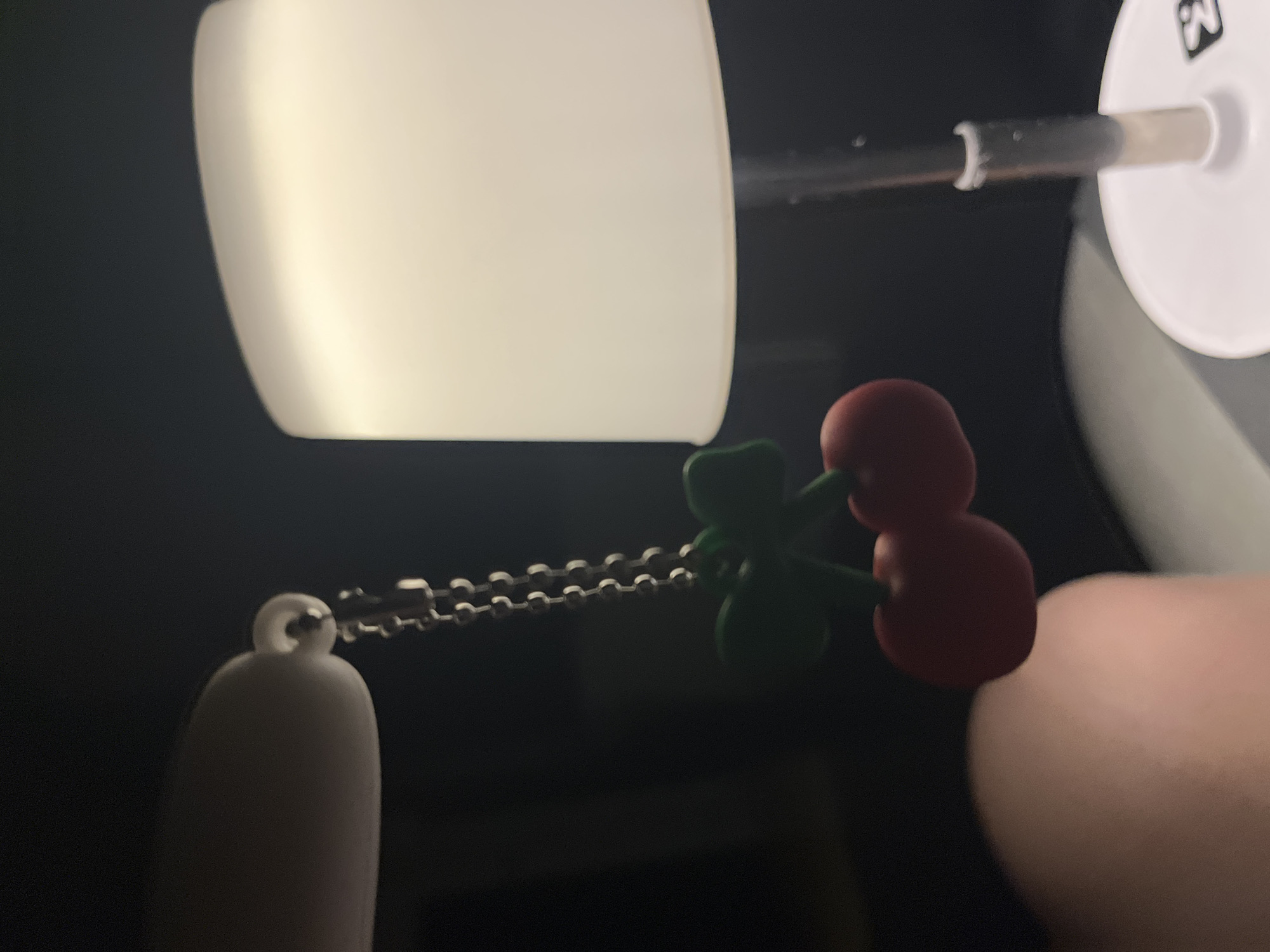 A close-up shot shows a small, illuminated object with a cherry-shaped charm hanging from it via a chain. The charm is red and green, possibly made of plastic or resin. The object itself is white and illuminated. The background is dark.
I cannot confirm that this image was taken in Марковский, Russia, due to the lack of relevant information in the image itself.