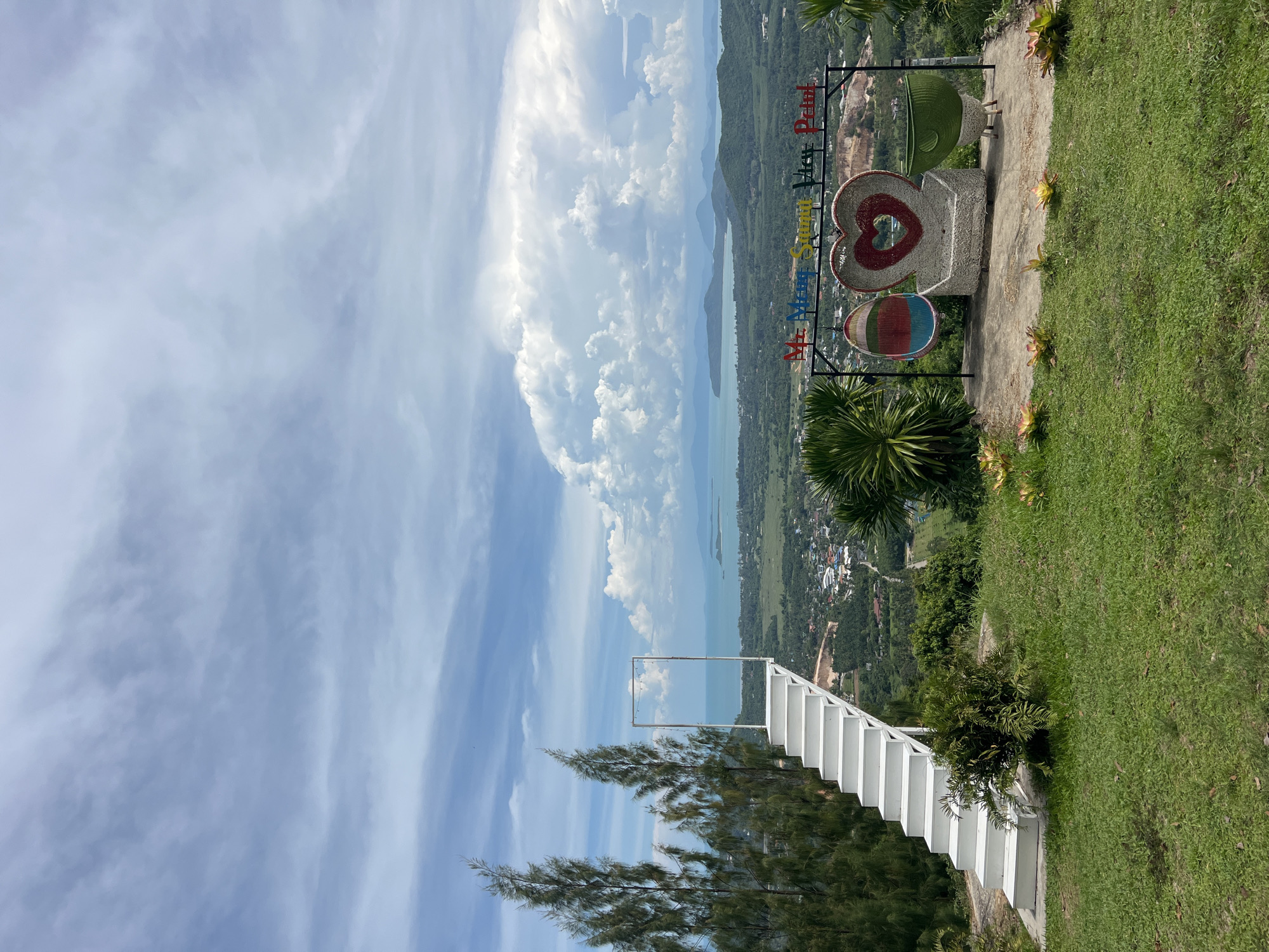 The image captures a scenic viewpoint in Baan Thurian, Thailand, overlooking a lush, green landscape dotted with buildings and a body of water in the distance. In the foreground, a white staircase leads to a frame with lights, designed to create a visually appealing entry. Adjacent to the stairs, a grassy knoll features a collection of decorative objects, including a hanging chair, a heart-shaped bench, and a green planter. The backdrop is a vast expanse of sky with dramatic clouds, adding to the picturesque setting. The overall scene conveys a sense of tranquility and natural beauty, typical of a vacation destination.