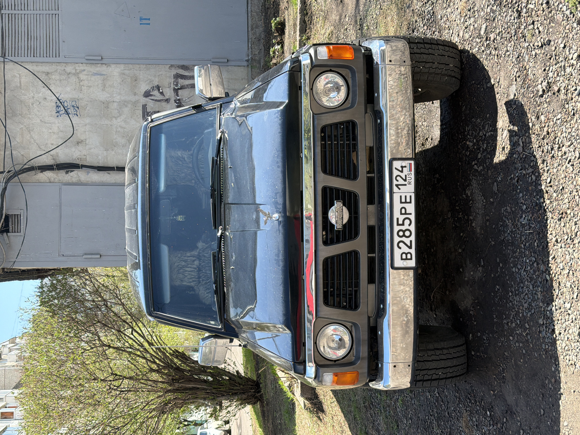 The image shows the front view of a dark-colored Nissan Patrol SUV, parked on a gravel and dirt shoulder next to a paved road in an urban environment. The vehicle is facing the viewer. The license plate reads "B285PE 124 RUS". The number "124" on the license plate indicates Krasnoyarsk Krai, Russia. Behind the vehicle is a light gray building with a large metal door and some graffiti on the wall. A tree with budding leaves is visible to the left of the SUV. The shadows indicate it is daytime.