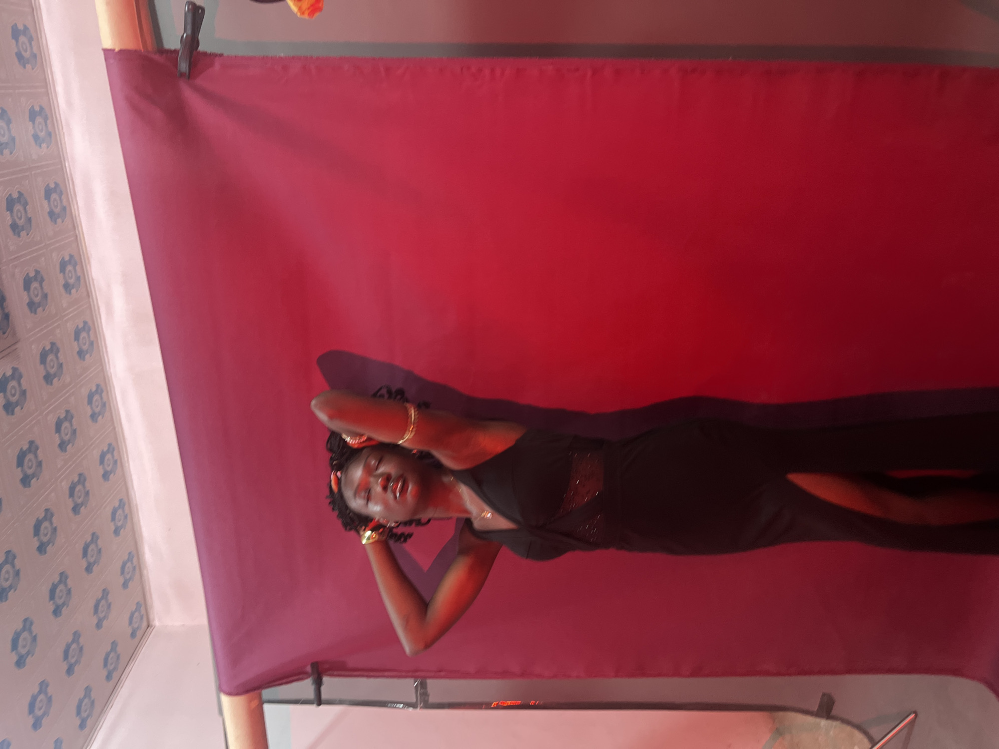 A young woman stands with her hands behind her head, eyes closed, in front of a large, maroon fabric backdrop. She is wearing a black sleeveless dress with a V-neckline and a thigh-high slit. Gold jewelry adorns her wrists and ears. The room has a white ceiling with a blue floral pattern. The lighting casts a red hue on the scene and creates a shadow of the woman on the backdrop. The overall impression is a posed studio portrait.