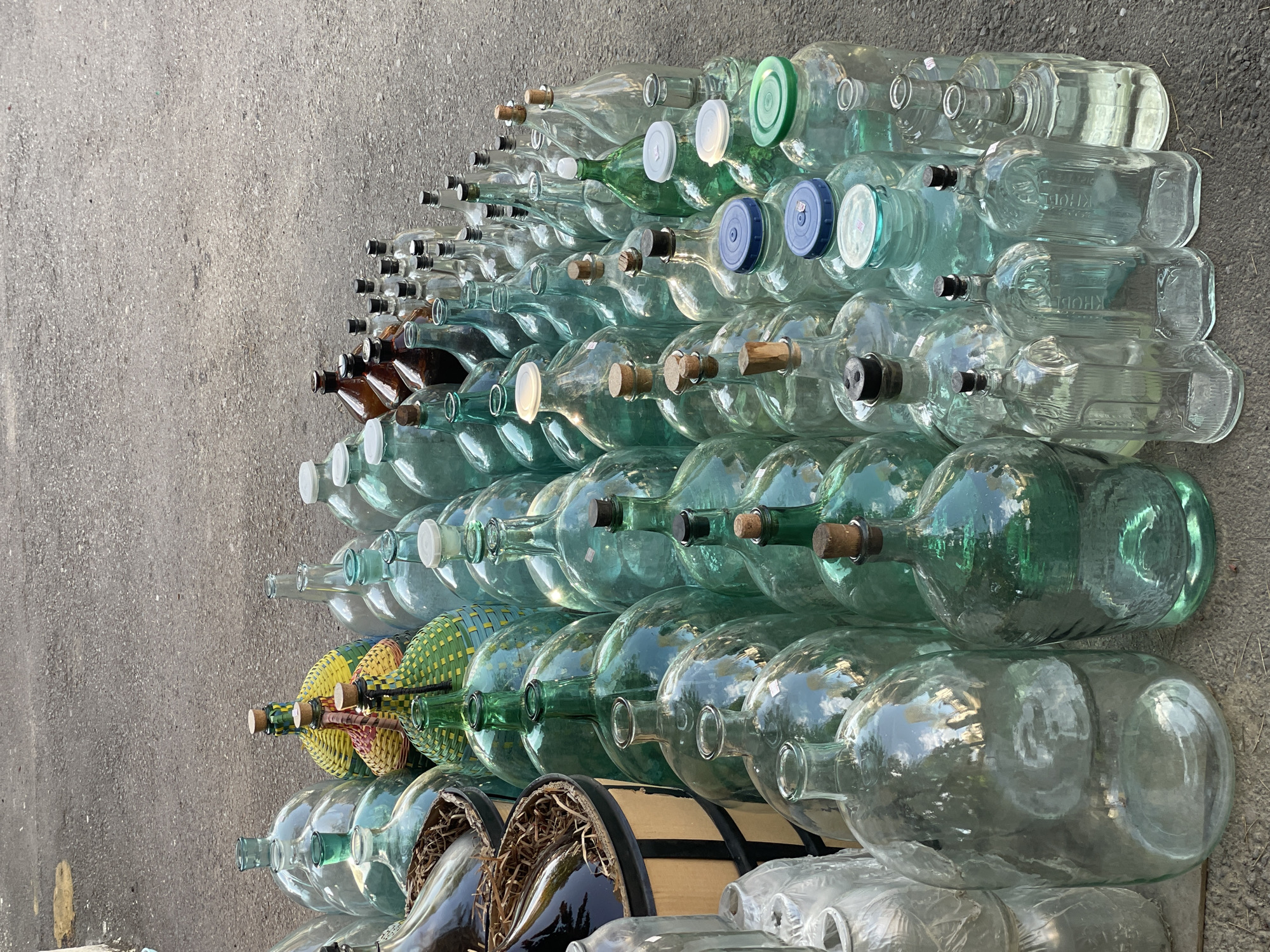 A large collection of glass bottles and carboys is stacked outdoors on a grey, paved surface, likely concrete or asphalt. The bottles vary significantly in size, shape, and color, with most being clear or light green, and a few amber-colored. They are arranged in multiple horizontal rows, forming a dense wall of glass that extends from the bottom right to the top left of the frame.
Many of the bottles have stoppers or caps. These include corks, dark black stoppers, and plastic caps in white, blue, and light green. Some of the larger, clear bottles appear to be carboys, and reflections of trees or foliage and the sky are visible on their curved surfaces, indicating bright daylight. Towards the center left, a few bottles are distinctively wrapped in colorful woven material, featuring patterns of yellow, green, and reddish-brown.
Visible embossed text on several clear bottles reads "KHOBE," with additional letters like "OLE" sometimes following, suggesting a brand or producer name such as "KHOBEOLE." The arrangement suggests the bottles are either being stored, awaiting recycling, or perhaps are part of a display or preparation for refilling, possibly related to local beverage production given the Tbilisi, Georgia context. The scene is brightly lit, indicating it is daytime. No people are visible.