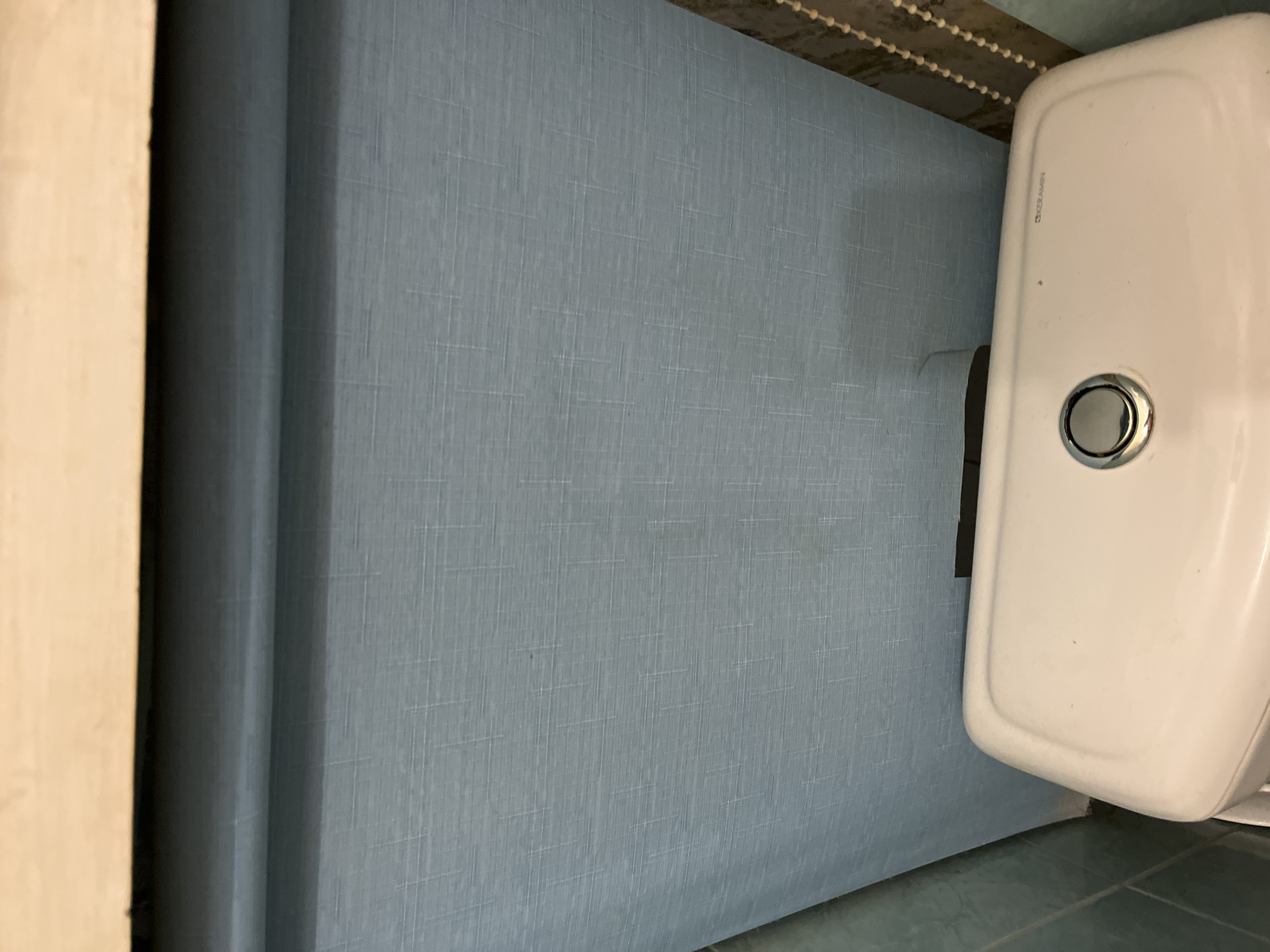 The scene depicts an overhead view of a bathroom interior in Minsk, Belarus. A light-colored wooden shelf or beam occupies the top portion of the frame. Below the shelf, a blue-grey textured wall covering, possibly wallpaper or a roller blind, extends vertically, displaying a linen-like weave pattern with subtle horizontal and vertical striations. A dark, irregular stain is present on the lower-center section of this wall covering.
At the bottom of the image, the top of a white ceramic toilet cistern is visible. On its left side, a circular chrome dual-flush button mechanism is centrally placed. Towards the right side of the cistern, the embossed brand name "KERAMIN" is clearly discernible. A narrow vertical strip of teal-colored tiled wall is partially visible on the far left. On the far right, a vertical string of small, light-colored spherical beads, consistent with a blind operating mechanism, is partially captured. No individuals, direct actions, or interactions are present in the image.