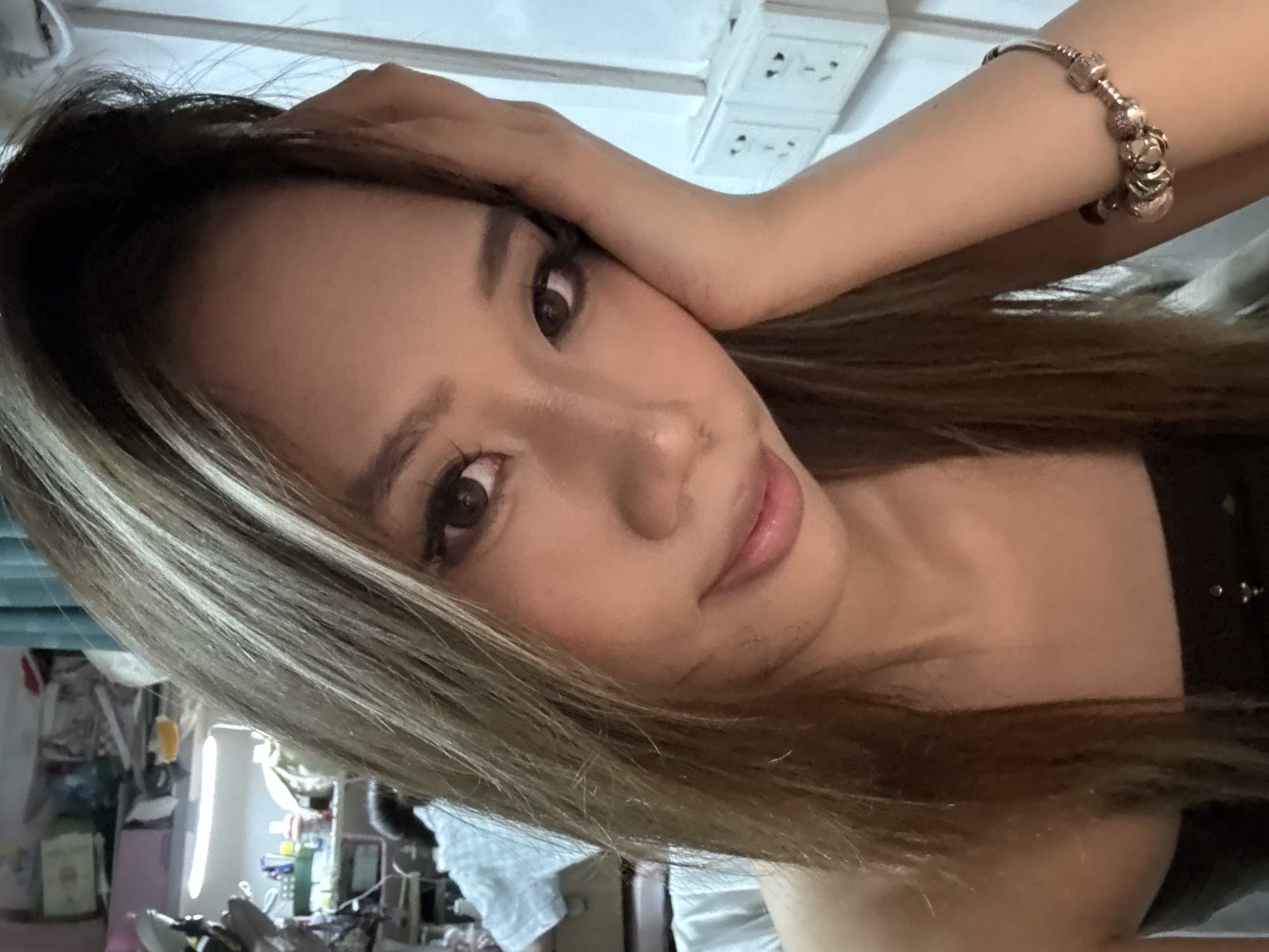 A young woman is holding her hand near her head and staring toward the camera. She is wearing a beaded bracelet on her right wrist. The background includes wall power outlets and a cluttered room. The location is suspected to be within the Nan'an District of China.