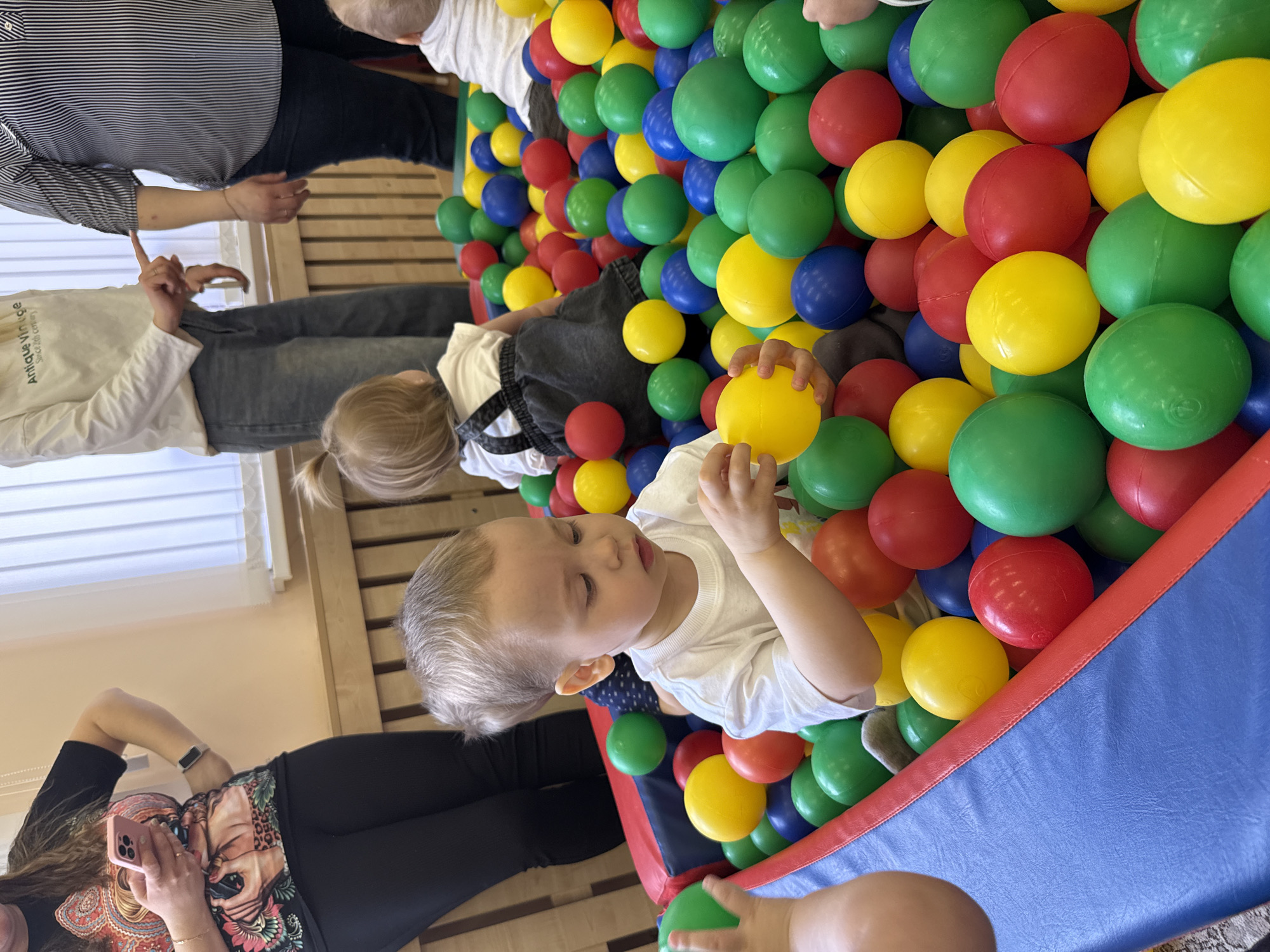 The image shows a group of people and children around a ball pit filled with multicolored plastic balls (yellow, green, red, and blue). A young child is sitting in the ball pit, holding a yellow ball and looking down. Another child with blonde hair is partially visible in the pit as well. Several adults are standing around the ball pit. One adult is holding a phone, likely taking the picture. The location is most likely an indoor play area. No specific details in the image confirm the Rusanovo location, however the presence of people indicates an inhabited area.