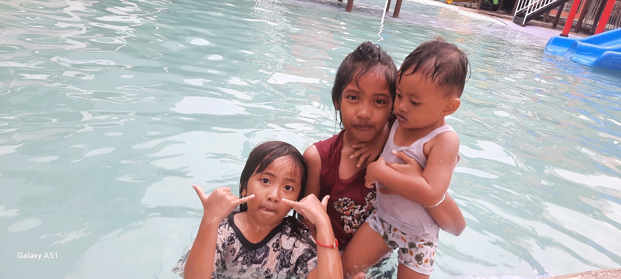 The image shows three children in a swimming pool. The child on the left is a young girl with dark hair, wearing a patterned top, and making a "shaka" hand gesture. The child in the middle is an older girl with dark hair pulled back, wearing a maroon sleeveless top. She is holding the child on the right, who appears to be a baby or toddler, wearing a light gray tank top and patterned shorts. They are all in clear, light blue water, with ripples on the surface. In the background, there are some structures and a blue slide, suggesting a water park or pool area. The bottom left corner of the image displays "Galaxy A51". The location is identified as Popoh, Indonesia.