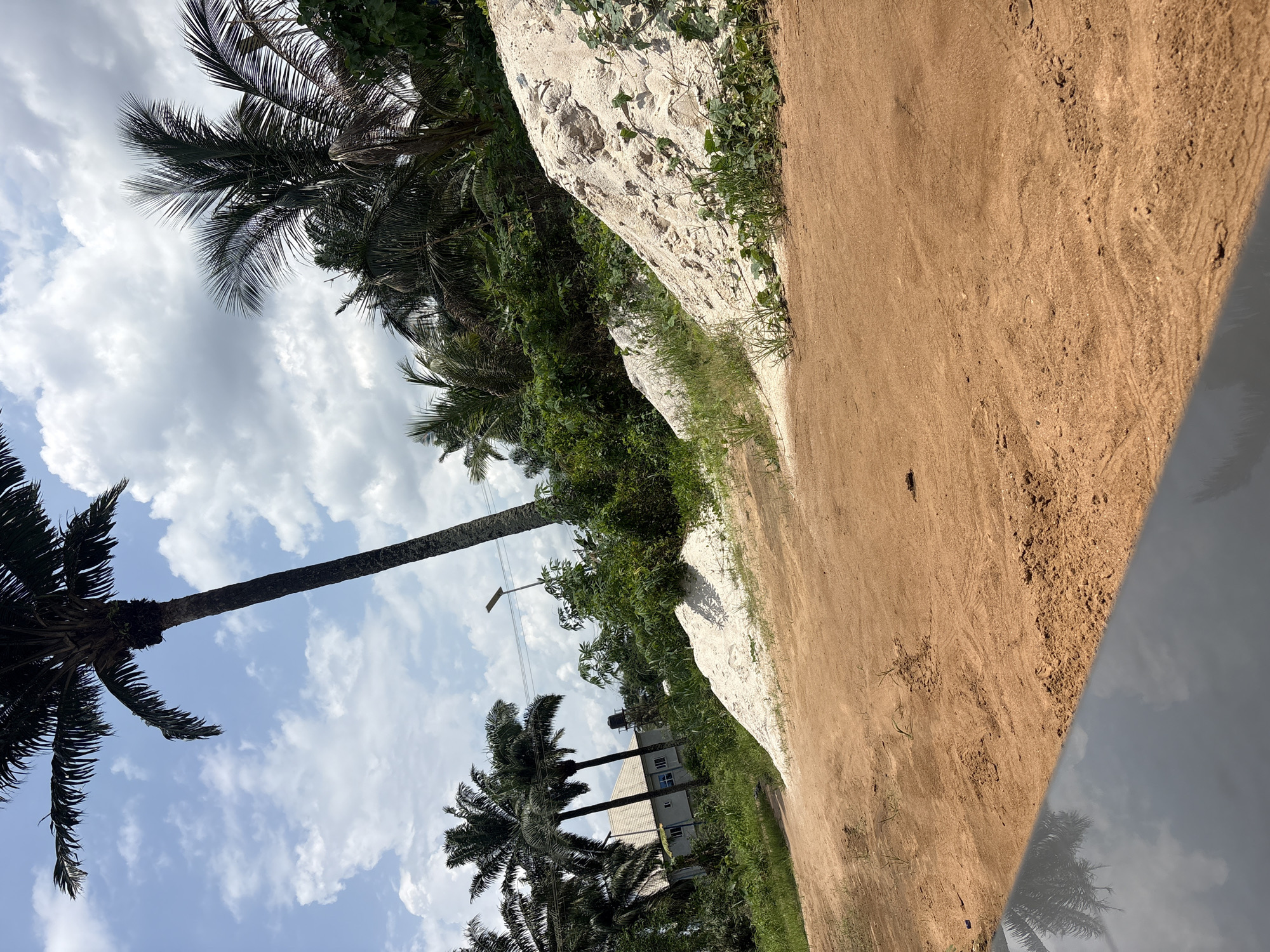 The image shows a dirt road in Ikot Ekpene, Nigeria, with a low, light-colored rock or concrete wall on one side, covered in vegetation. Tall palm trees line the opposite side of the road, reaching into a sky with scattered clouds. A building can be seen partially obscured by the trees. The image appears to have been taken from a vehicle, as part of the vehicle's body is visible in the lower right corner.