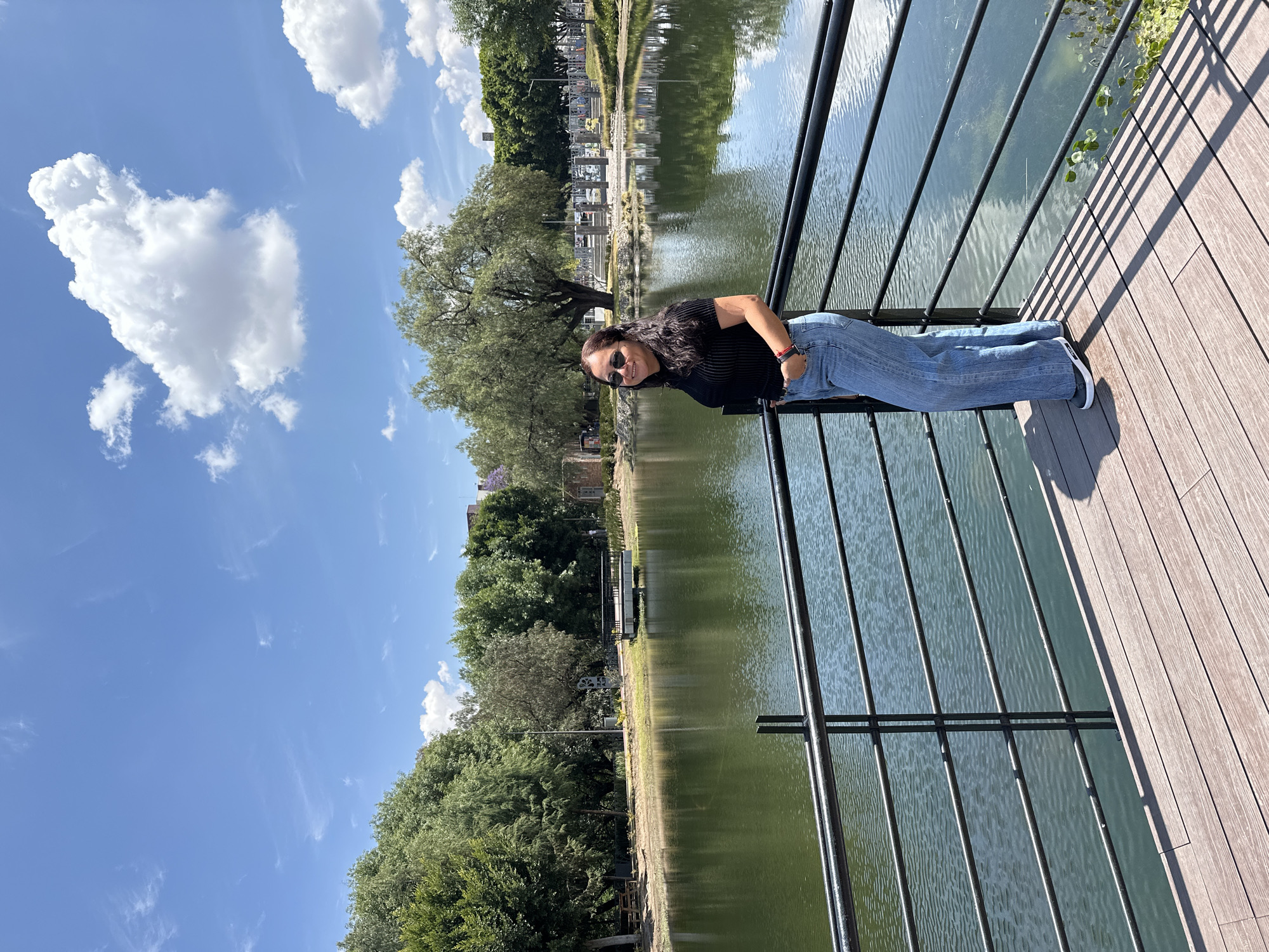 The image shows a woman standing on a wooden boardwalk next to a lake, seemingly leaning against the railing. The woman is wearing sunglasses, a black shirt, and blue jeans. She has her left hand in her pocket and is smiling slightly. The backdrop features a body of water with reflections of trees and buildings. The sky is blue with fluffy white clouds. The image appears to be taken in Puebla, Mexico.