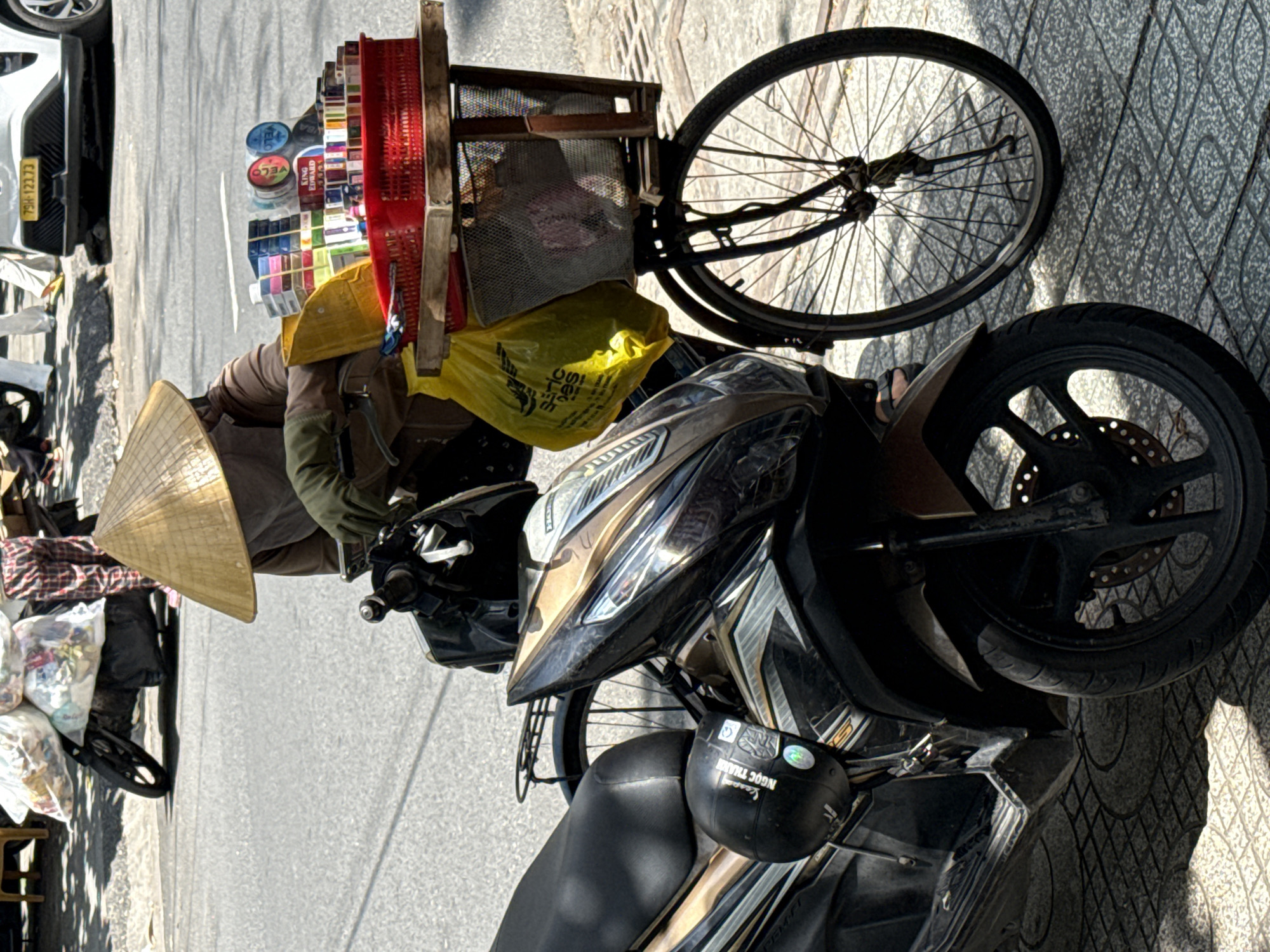 In Nha Trang, Vietnam, a vendor wearing a conical hat stands beside a parked motorcycle. The vendor is on a bicycle that is loaded with various items, including cigarette packs, small cosmetic products, and a yellow plastic bag. The motorcycle is parked next to the bicycle, and the scene is set on a paved sidewalk next to a road. The vendor appears to be selling goods from their bicycle, as indicated by the display of products. The environment suggests a typical street scene in Vietnam, with the presence of both a motorcycle and a bicycle, common modes of transportation in the region.