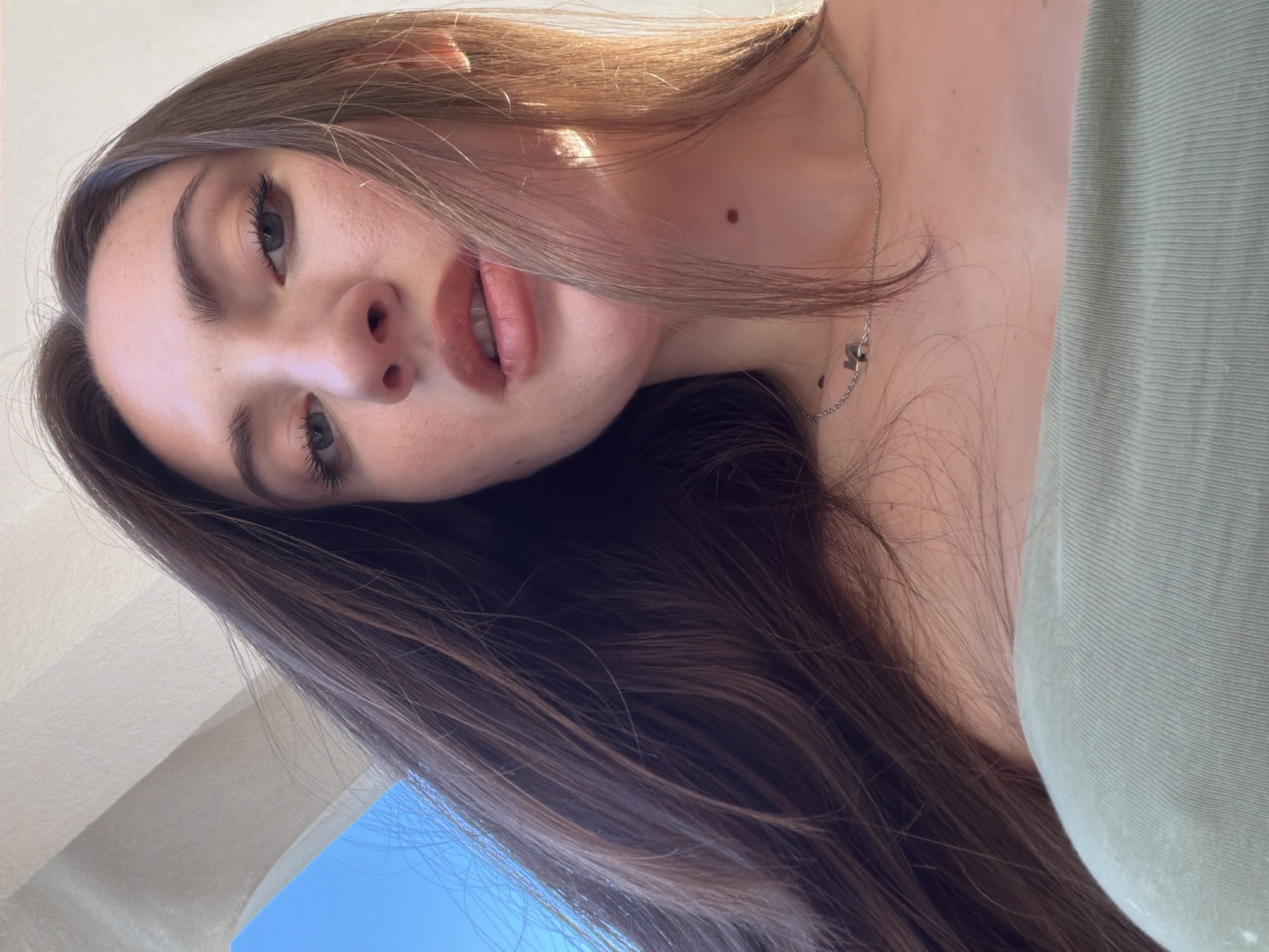 A low-angle, close-up, outdoor, sunlit selfie of a young woman with long, dark brown hair. She is wearing a light green, off-the-shoulder top and a delicate silver chain with a letter pendant. Her face is turned towards the camera, and her gaze is directed slightly upwards. She has natural makeup, with defined eyebrows, mascara, and a light pink lipstick. A small, dark mole is visible on her neck. The background shows a bright blue sky and a cream-colored architectural element, possibly a canopy or awning, suggesting an outdoor setting in Sharm El Sheikh, Egypt.