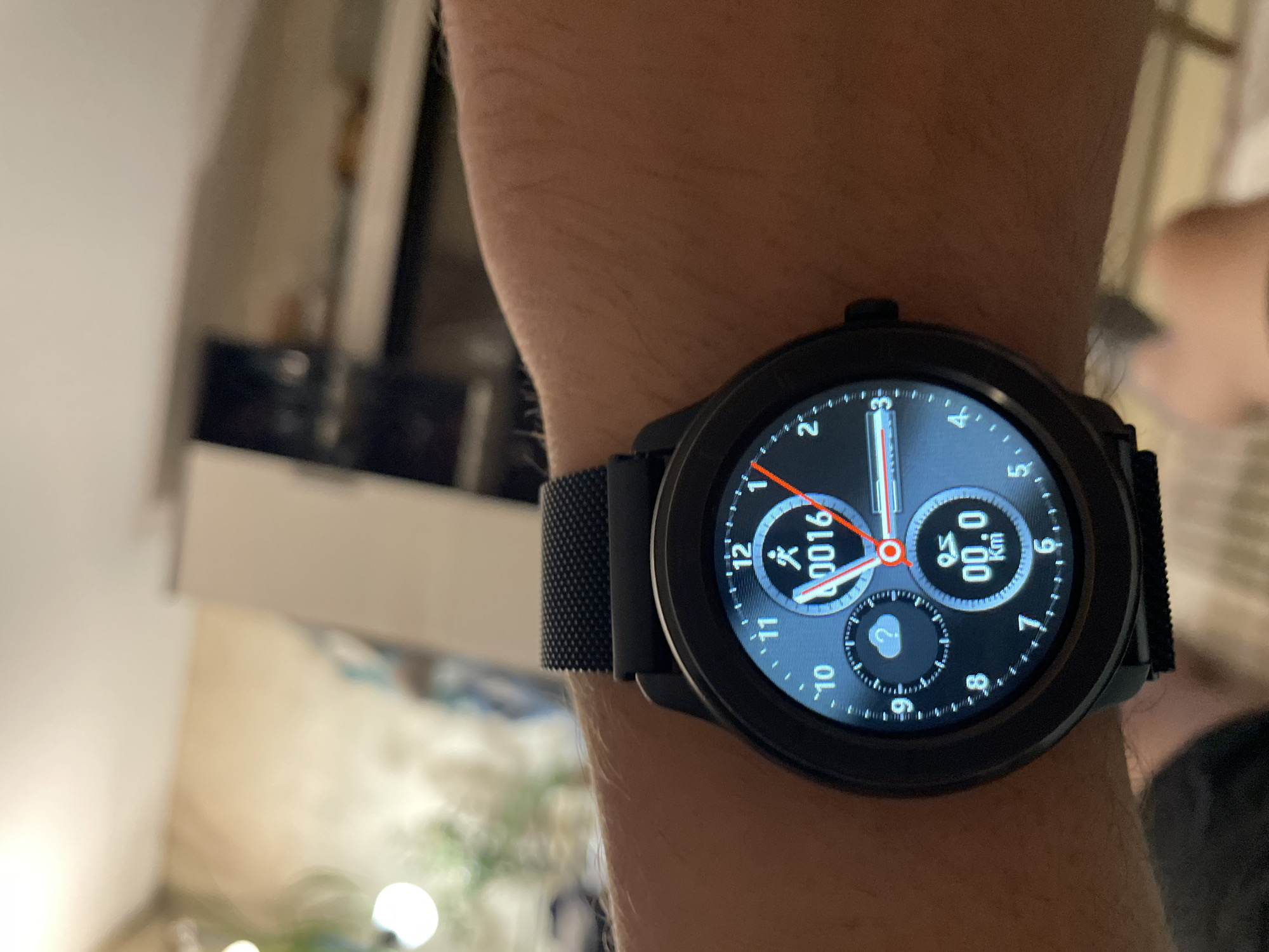 A close-up, eye-level shot shows a person's wrist wearing a black smart watch with a round, black bezel and a black woven band. The watch face displays a digital interface with a blue background. At the top, the number "12" is visible, with a runner icon next to the number "0016". Below this, a smaller circular dial shows a cloud icon with a question mark inside. Further down, another circular dial displays "00.0" with "Km" underneath. The watch hands are red and white, with the hour hand pointing to approximately 1:30 and the minute hand pointing to approximately 5:30. The background is blurred, showing what appears to be an indoor setting with furniture and lighting. There is no information in the image to suggest the location is Ufa, Russia.