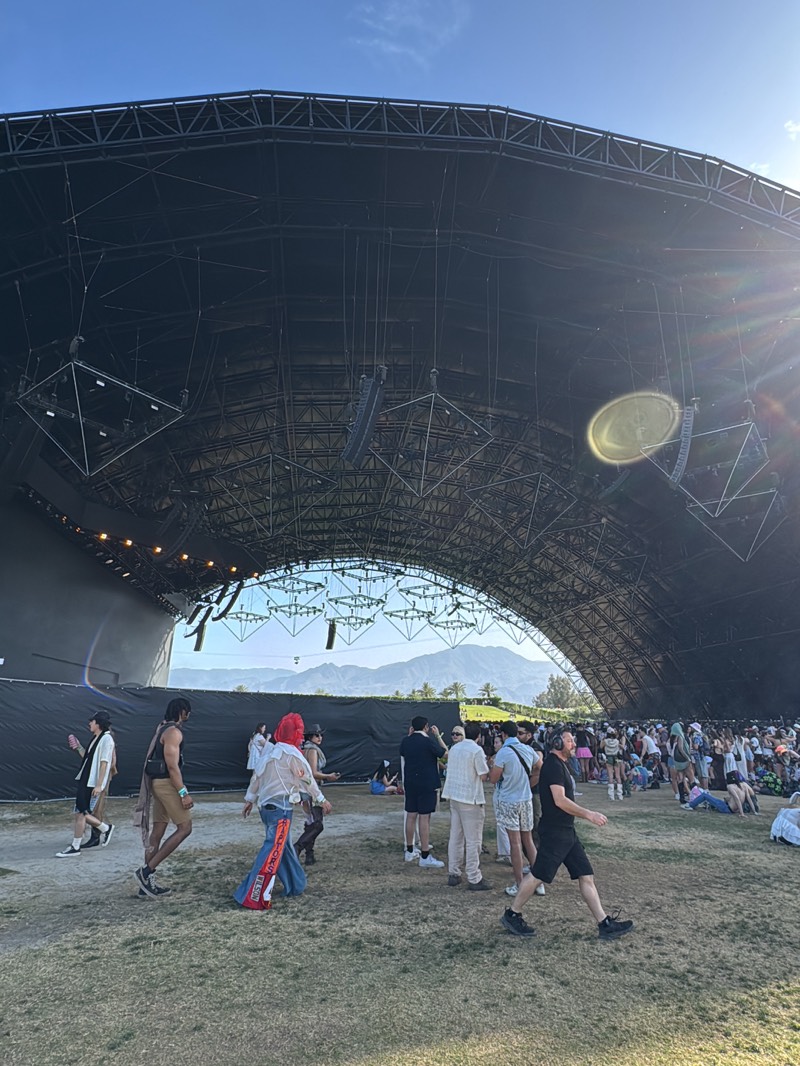The image shows a crowd of people gathered outdoors under a large, arched structure. The structure appears to be a stage or performance area, with rigging and lighting equipment hanging from its ceiling. In the background, mountains are visible. The people are dressed in a variety of casual clothing, and some are walking around while others are sitting or standing. The ground is covered in grass. The scene is lit by natural daylight, with a bright sun shining in the upper right corner of the frame. The location appears to be Palm Desert, United States, based on the desert environment and mountainous backdrop, consistent with the Coachella Music Festival.