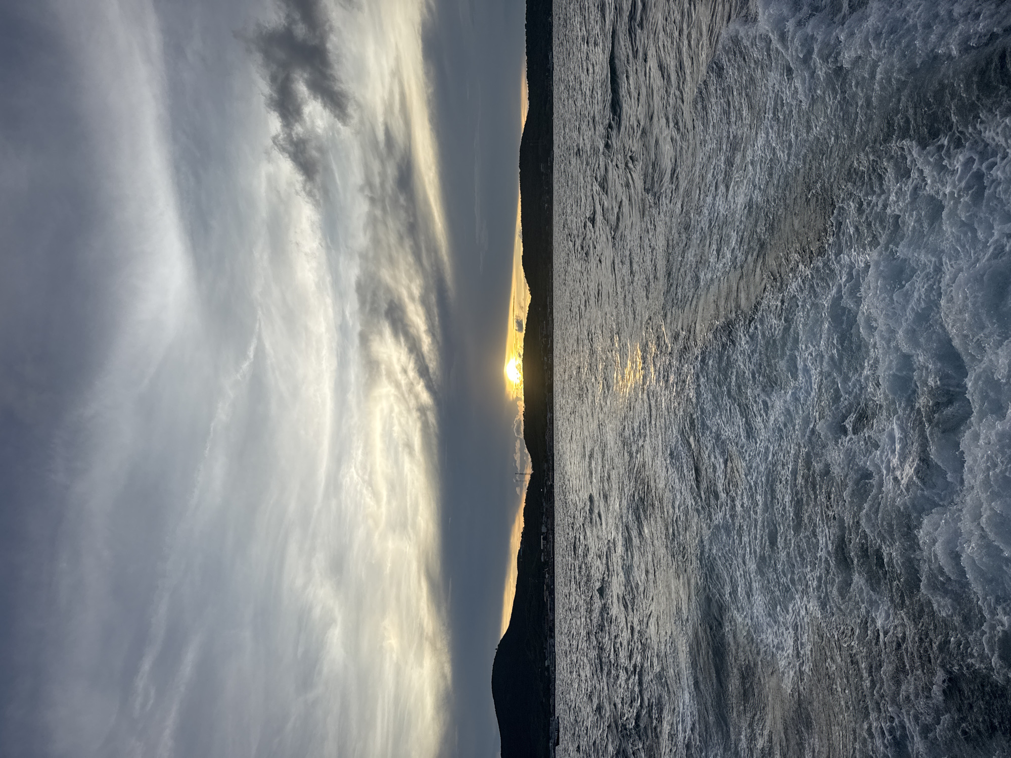 The image depicts a sunset scene in Pattaya City, Thailand. The sun is setting over a body of water, likely the Gulf of Thailand, casting a golden reflection on the waves. Silhouetted hills or landmasses are visible in the distance. The sky features striated clouds, some gray and others illuminated by the setting sun. The foreground shows turbulent water with whitecaps, indicating waves or strong currents. No people or specific objects are clearly discernible beyond the natural elements of the landscape.