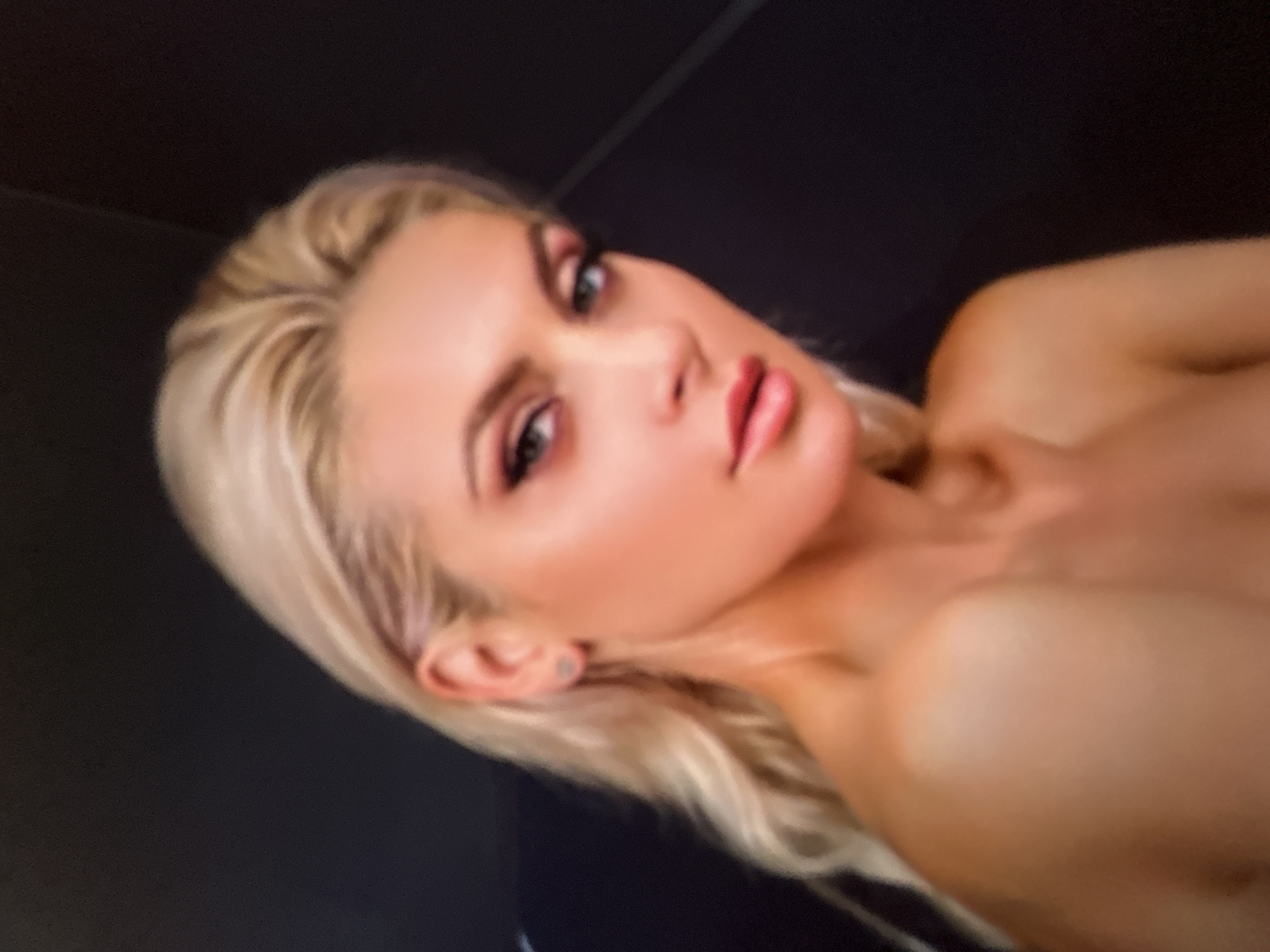 The media file is a close-up, low-angle shot of a blonde woman with fair skin and light makeup. She is looking towards the camera with a slightly parted mouth. Her hair is a medium blonde and falls around her shoulders. She is wearing what appears to be a dark top, and a portion of her upper chest is visible. The background is dark and out of focus, providing no discernible details about the setting. The lighting is soft, highlighting her features. The image appears to be taken indoors, possibly in a dimly lit room, with no indication of the time of day or weather conditions. There is no visible text in the image.