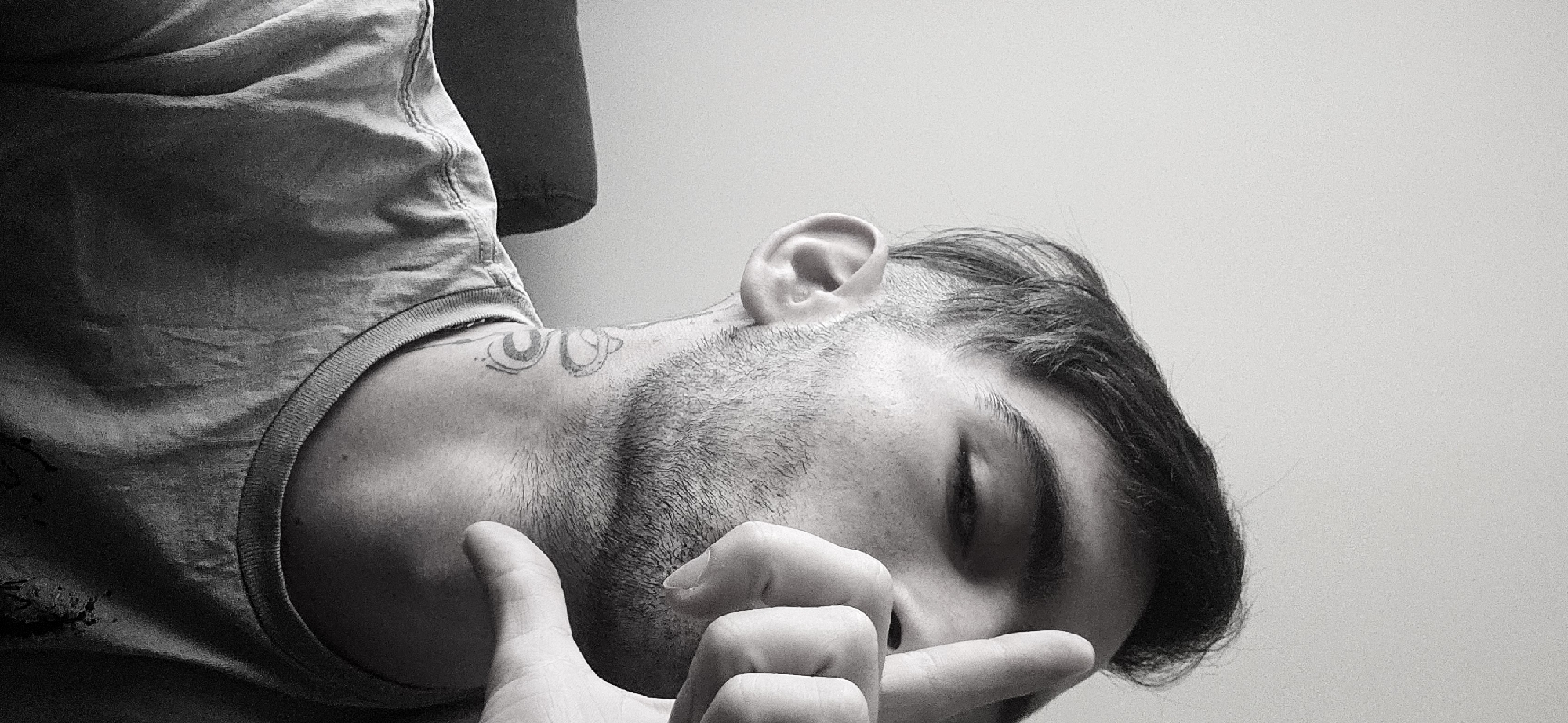 A monochrome, horizontally oriented photograph presents a close-up of a person, likely male, captured in a moment of rest or contemplation. The individual is positioned with their head tilted upwards and to the right, implying they are lying down or leaning back against a surface. Their eyes are closed, and one hand covers the lower portion of their face, obscuring the mouth and nose.
The subject has short, dark hair, styled with longer strands on top and closely cut or faded sides, exposing the ear. A visible stubble covers their jawline and extends onto their neck. A distinctive, partially visible tattoo, consisting of what appears to be two stylized, interconnected or mirrored shapes, is located on their left shoulder/collarbone area. They are wearing a plain, light-colored t-shirt with a ribbed collar, the fabric showing slight wrinkles and texture.
The background is a simple, light-toned wall, softly blurred, suggesting an indoor environment. The lighting is gentle and diffused, creating a serene atmosphere and highlighting the textures of skin and fabric. The black and white tonality adds to the pensive or intimate mood of the scene. No discernible text is visible within the image. While the image itself provides no direct geographical cues, the context states the location is Plovdiv, Bulgaria.