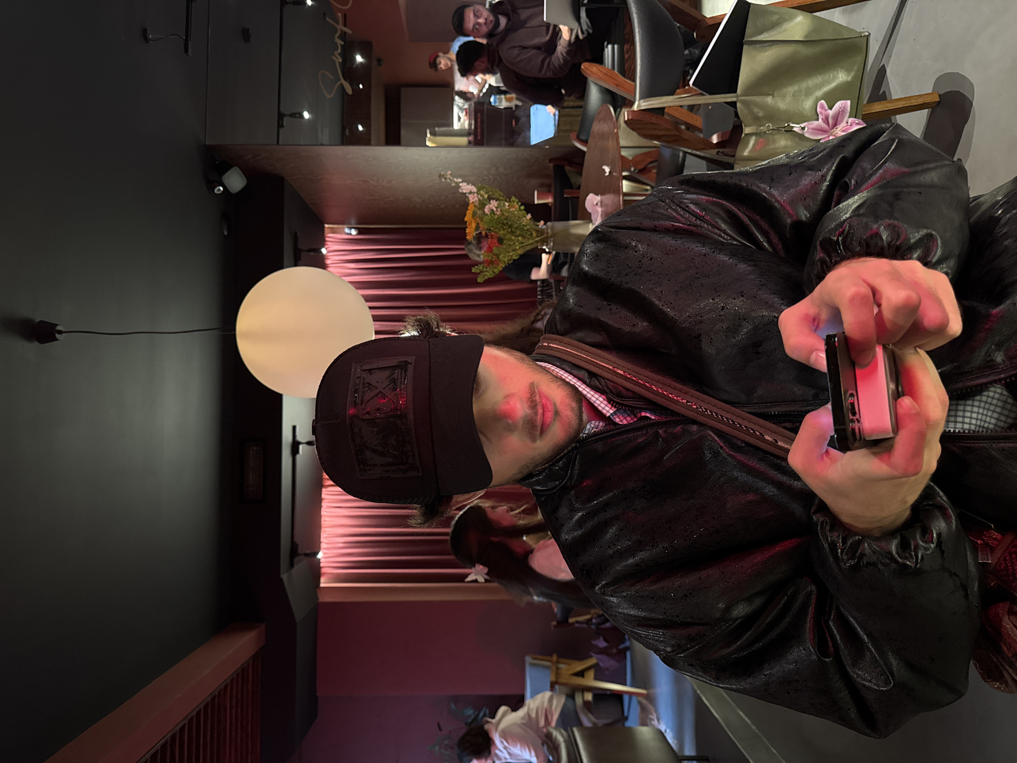 A medium shot depicts a person holding a mobile device, likely a smartphone, in a dimly lit indoor environment. The person is wearing a dark, textured jacket, and a dark baseball cap with a pattern on the front. They are positioned at an angle, facing slightly towards the camera, with their right hand holding the device. The background suggests a restaurant or bar setting, with tables, chairs, and other patrons visible in the distance. A white globe-shaped light fixture is noticeable on the left side of the frame, and red curtains or drapes are behind the subject. The exact location in Yerevan, Armenia, cannot be determined from the image alone.