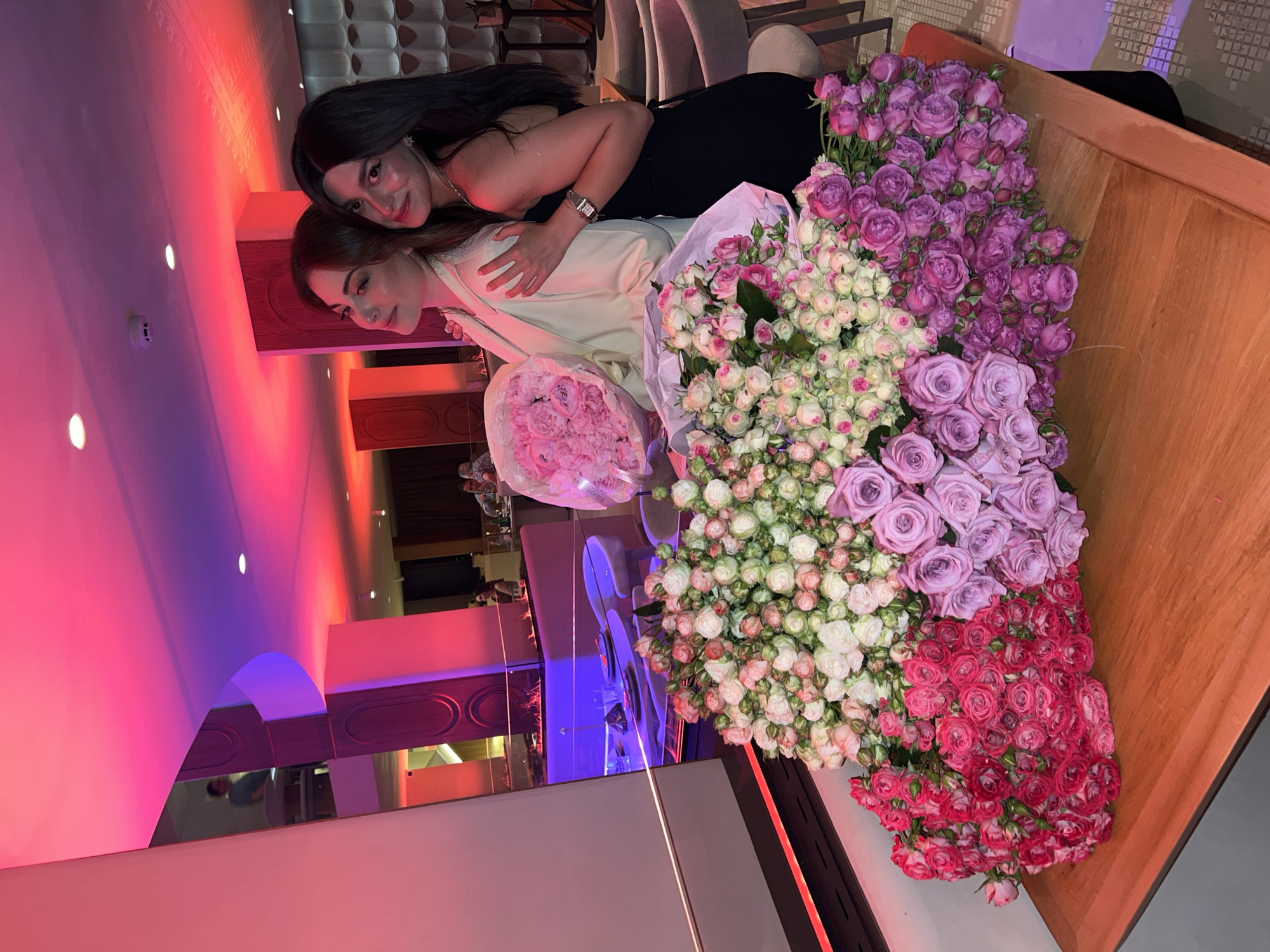 Two women are posing with several bouquets of roses, including light pink, white and light pink, light purple, and red roses. One woman is wearing a black dress and a wristwatch. The other woman is wearing a white jacket. The setting appears to be an upscale indoor location. The background includes architectural details illuminated with red and blue lighting. Yerevan, Armenia is not evident in the image.