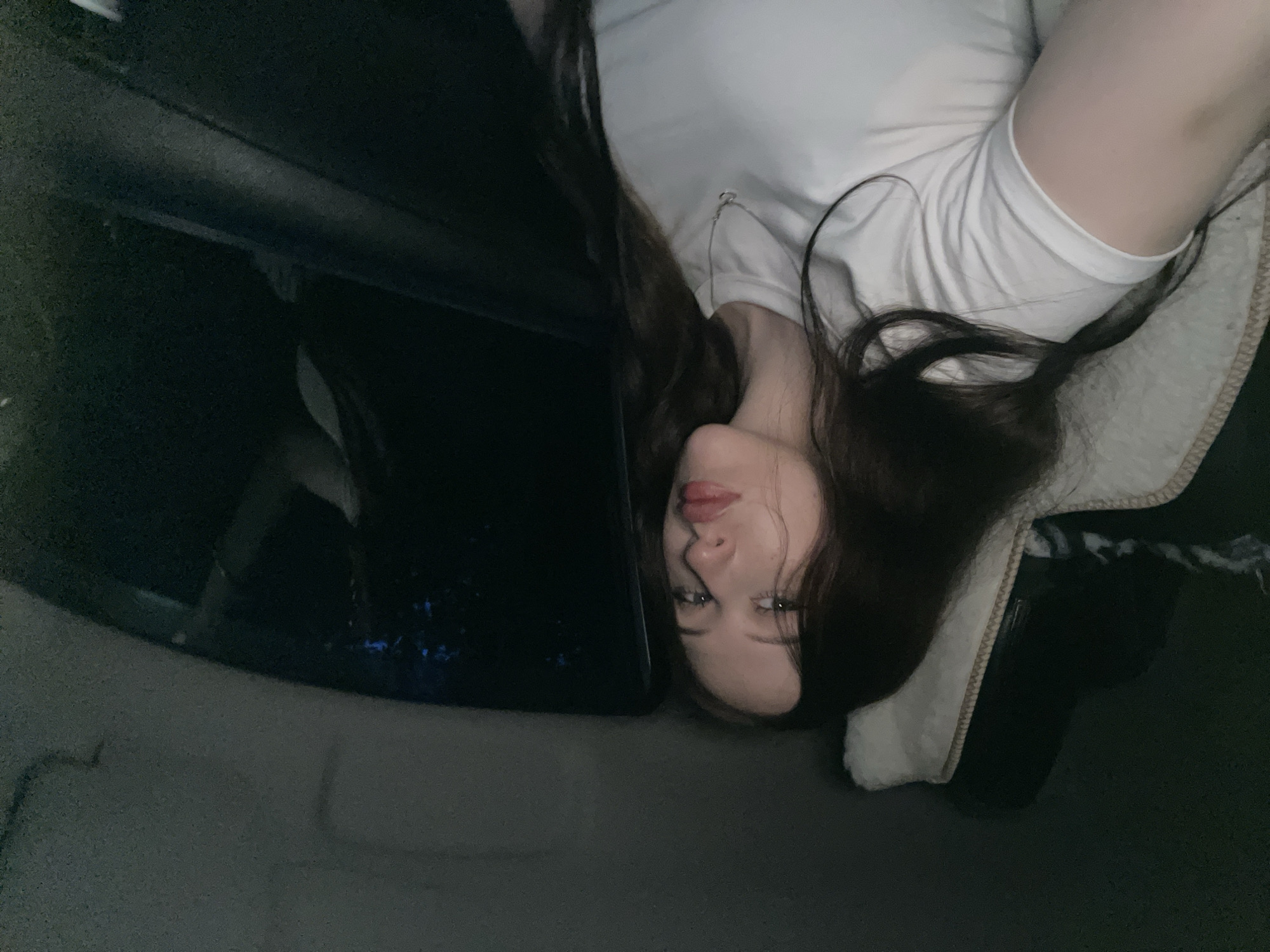 A young woman with long dark hair and light skin is sitting in a car at night, looking at the camera. She is wearing a white t-shirt and a silver necklace. The car interior is dimly lit, with the exterior appearing dark and possibly rural. Her face is turned slightly to the left, and her lips are slightly parted. The background shows the dark interior of the car, including a window with a faint reflection and what appears to be a headrest covered with a blanket.