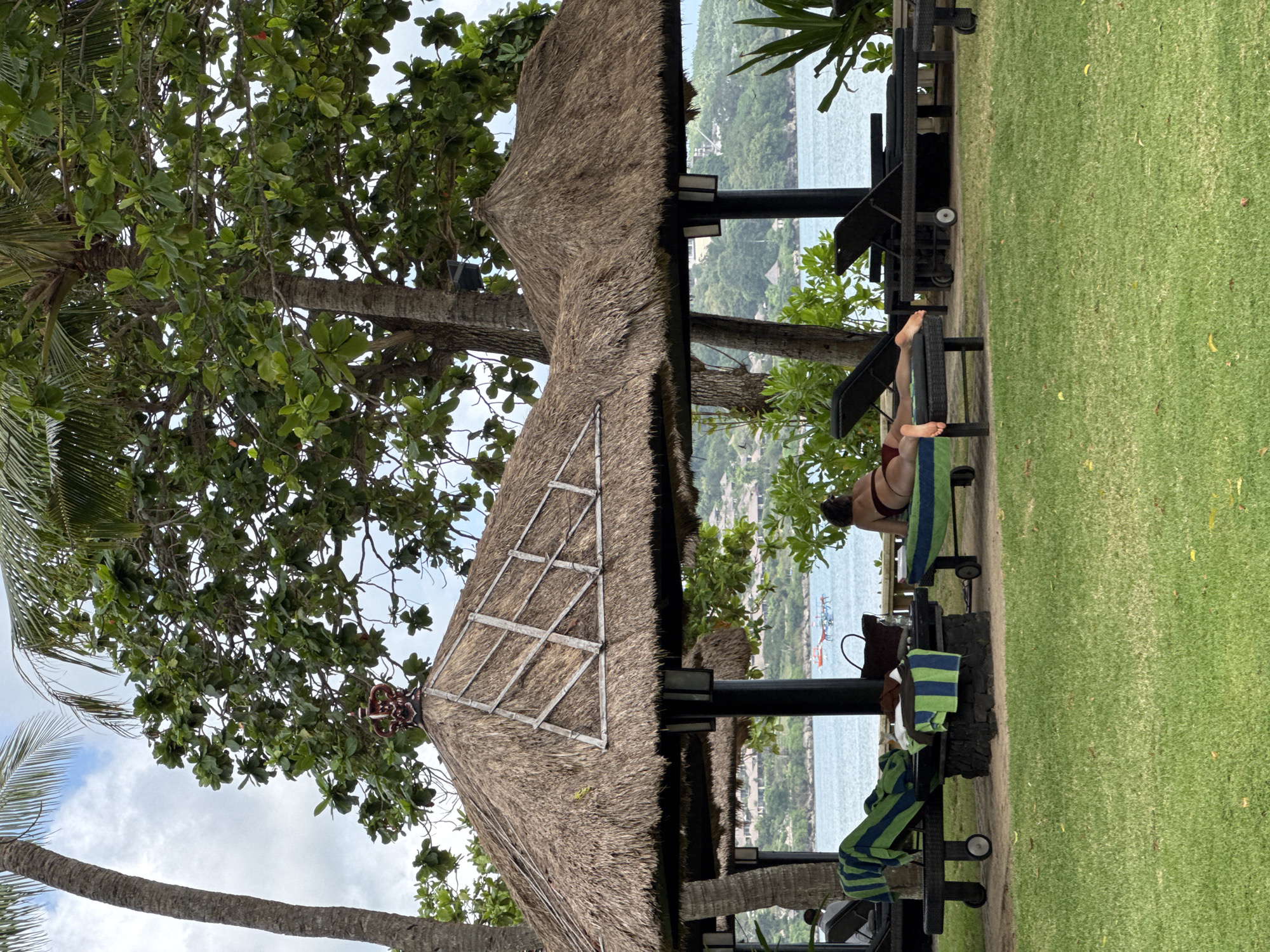 The scene is located in Kuta Selatan, Indonesia. A thatched-roof structure stands on a well-manicured lawn. Underneath, a person reclines on a lounge chair near the ocean. Towels and bags are nearby. Trees with dense foliage surround the structure. In the distance, the ocean meets the coastline. The sky is partially cloudy.