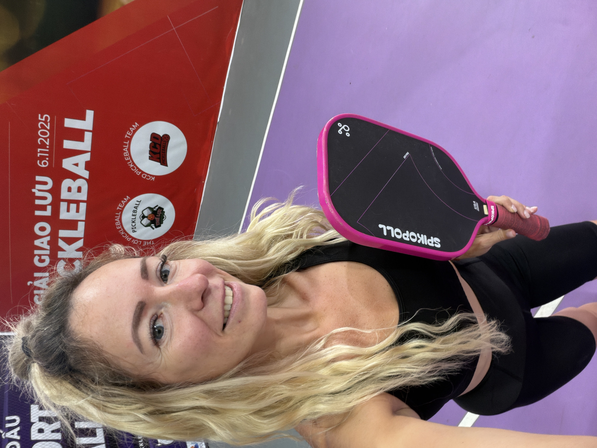 The image shows a blonde woman with her hair in a high ponytail, wearing a black sports bra and black cycling shorts, holding a pink pickleball paddle. She is looking towards the camera with a slight smile. Behind her, a red banner with white text reads "GIẢI GIAO LƯU 6.11.2025 PICKLEBALL". The background also shows a purple court surface and a portion of a gray wall. The event appears to be a pickleball tournament or exchange held in Da Lat, Vietnam, on November 6, 2025.