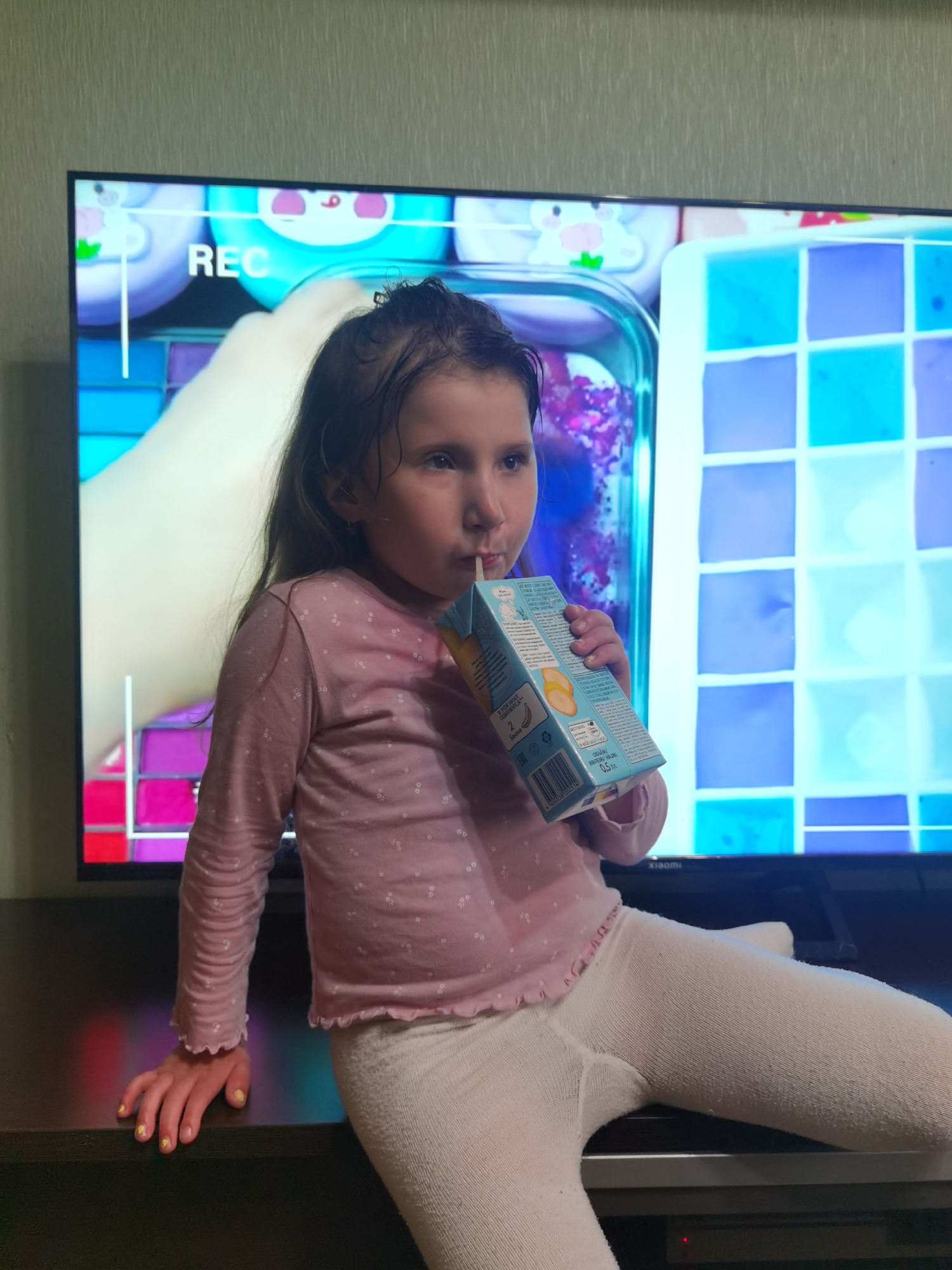 The scene is an indoor setting in the city of Chaikovsky, Russia. A young girl, approximately 5-8 years old, is seated on a dark brown, flat-surfaced piece of furniture positioned in front of a large, flat-screen television. The girl has long, dark hair, which appears slightly unkempt. She is dressed in a light pink long-sleeved shirt adorned with small white patterns and light-colored tights. She holds a light blue and yellow juice box, marked with Cyrillic text, a barcode, and indicating a volume of 0.5 liters, from which she drinks via a white straw. Her left hand rests on the dark furniture surface; her fingernails are painted yellow with small, subtle designs. Her attention is directed toward the television screen, which displays vibrant animated content, including a light-skinned hand, various containers, and a grid of blue and purple squares. The letters "REC" are visible in the upper left of the screen, and a "Xiaomi" logo is discernible on the television's lower bezel. The wall behind the television is covered with light-colored, vertically textured wallpaper.