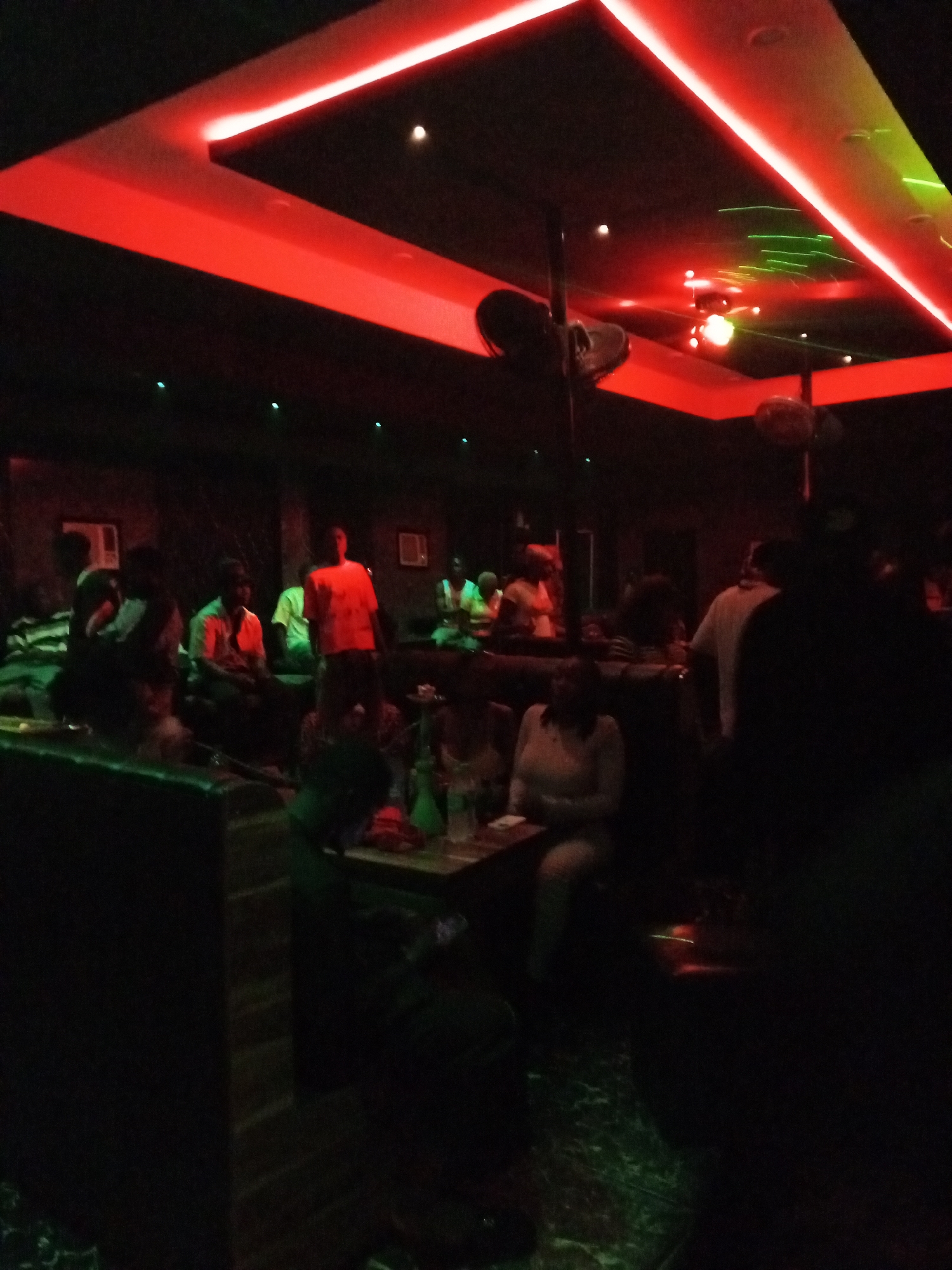The image captures a dimly lit indoor scene, likely a club, lounge, or bar, set in Lokoja, Nigeria, during nighttime. The space is characterized by its dark walls and ceiling, dramatically illuminated by vibrant red neon strip lighting that forms geometric patterns on the ceiling. Green laser lights sweep across the upper part of the room, adding to the atmospheric lighting.
Multiple people are present, socializing in small groups. Some individuals are seated at tables in booth-style or upholstered chairs, while others appear to be standing or moving around. Due to the low light conditions and distance, individual faces and precise details of their attire are mostly indistinct, appearing as silhouettes or partially illuminated figures. One person in the mid-ground wears a brightly lit orange shirt, drawing the eye, and another in the foreground is visible in a light-colored top.
Objects include several tables, at least one of which prominently displays a shisha or hookah pipe with its characteristic hoses, indicating a shisha lounge environment. White wall-mounted units, possibly air conditioners or light fixtures, are visible on the dark walls in the background. Overhead, there are what appear to be dark, hanging speakers or decorative elements. The floor has a subtle reflective quality, possibly tiles, with hints of green light. No visible text is discernible in the image. The overall impression is one of a lively, yet relaxed social gathering space.