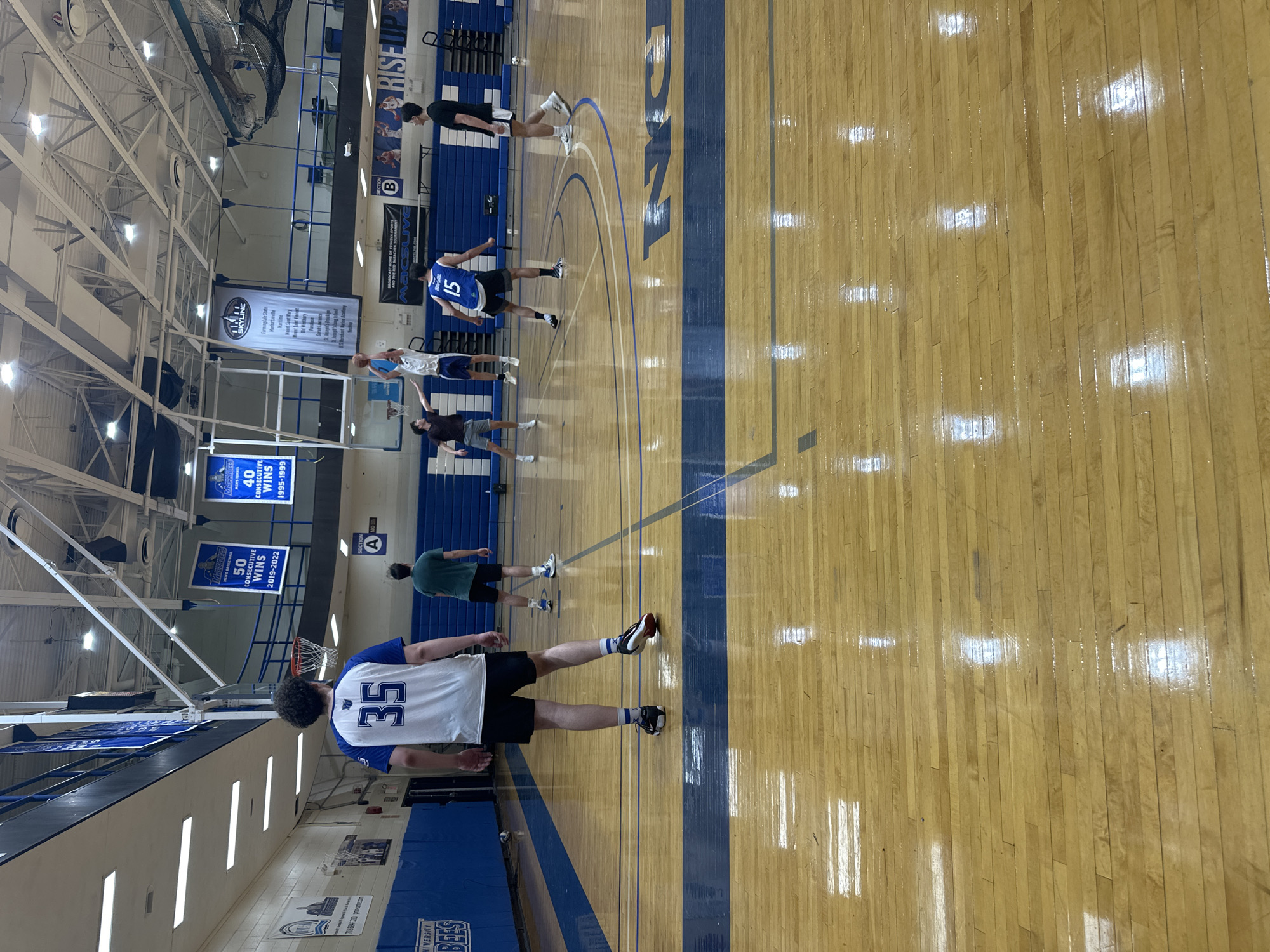 The image captures an indoor basketball court within a brightly lit gymnasium, likely at a college or university. The wooden floor features distinct blue and white lines, along with the blue painted letters "FSC" and the word "FALCONS," indicating the institution's abbreviation and mascot. The high ceiling exposes metal trusses and is illuminated by numerous fluorescent light fixtures. Blue bleachers are visible along one side of the court, and basketball hoops are positioned at either end.
Several males, appearing to be young adults or college athletes, are scattered across the court, dressed in athletic wear. They seem to be participating in a basketball practice session or warm-up. In the foreground, a person in a white jersey with blue trim and the number "35" on the back stands facing away from the camera. Further down the court, another individual wears a blue jersey with the number "15." Other participants are dressed in t-shirts and shorts.
Banners and signs hanging from the walls and ceiling provide notable details. Text visible includes "SKYLINE," "Farmingdale State," and "Maritime," suggesting affiliation with a conference or specific colleges. Achievement banners read "MEN'S TENNIS 40 CONSECUTIVE WINS 1995-1999" and "50 CONSECUTIVE WINS 2019-2022." Other visible text includes "ABCSLIVE.COM," "RISE UP," "SECTION A," and "SECTION B." Given the "Farmingdale State" references and the New York context, the location is likely Farmingdale State College in New York, USA.