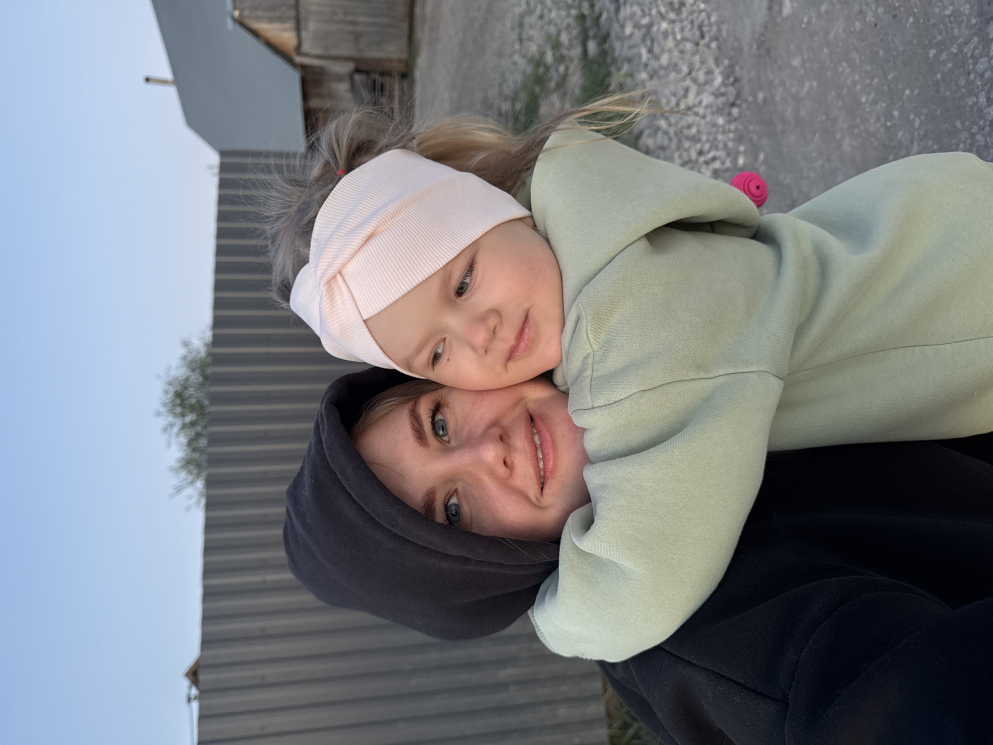 A selfie-style photograph depicts a fair-skinned woman with blue eyes, dressed in a dark hooded sweatshirt, embracing a young child with blonde hair. The child wears a light pink headband and a pale green hooded sweatshirt. The pair are outdoors; behind them are buildings and a gravelly ground surface. The photograph was taken in Горный, Russia.