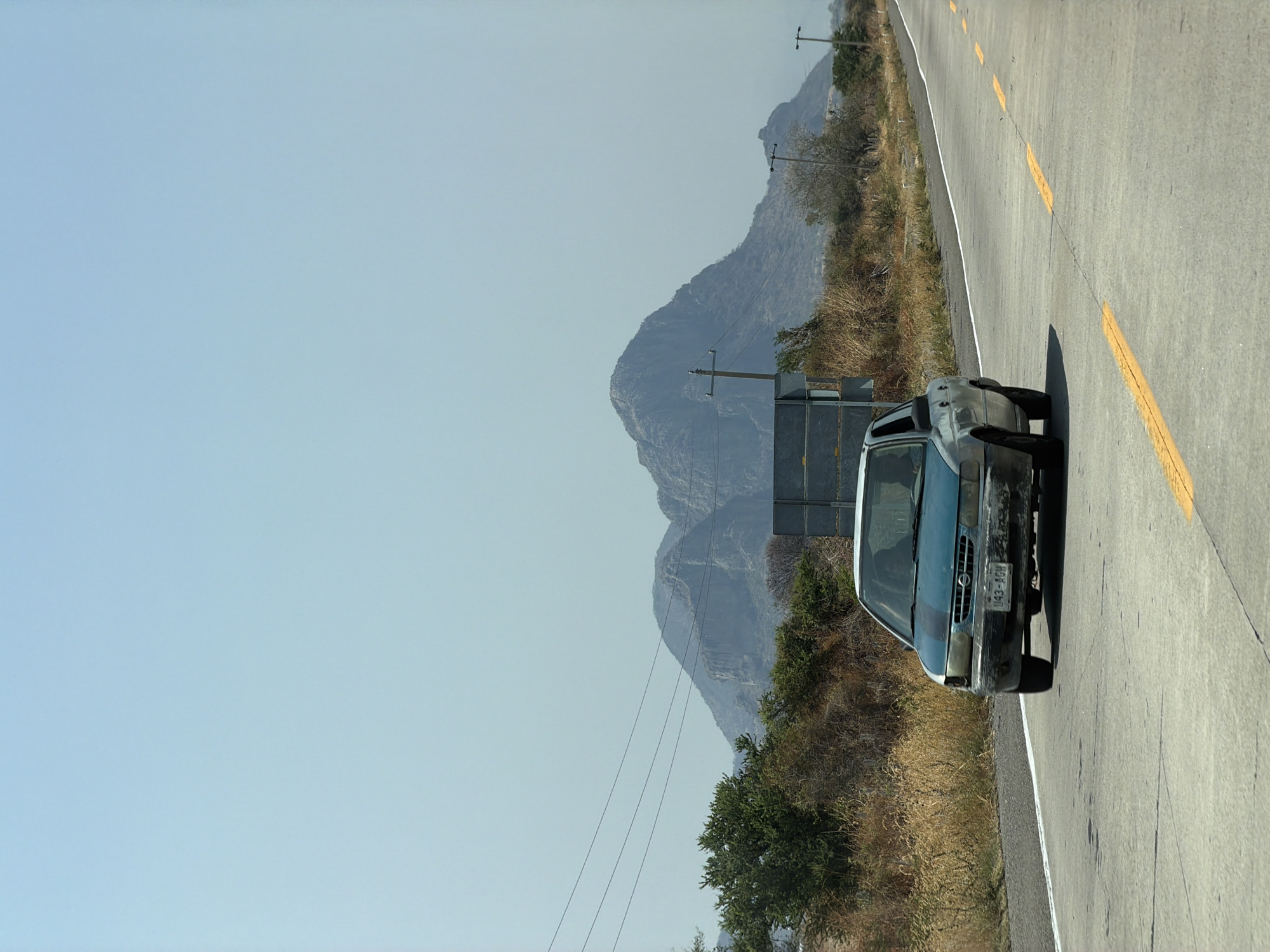 A horizontal, eye-level shot depicts a highway in Jantetelco, Mexico. The image is composed of a car parked on the side of the road with a mountain range and a clear sky in the background. The highway, made of gray concrete, features yellow dividing lines on one side and a white line on the other. Adjacent to the road is a grassy area interspersed with shrubs. The vehicle, a silver-blue Nissan sedan, is parked off the side of the road, facing towards the camera. Behind the car, a utility pole and sign are visible. The mountain range in the background is characterized by rocky peaks. The sky above is a clear, light blue color.