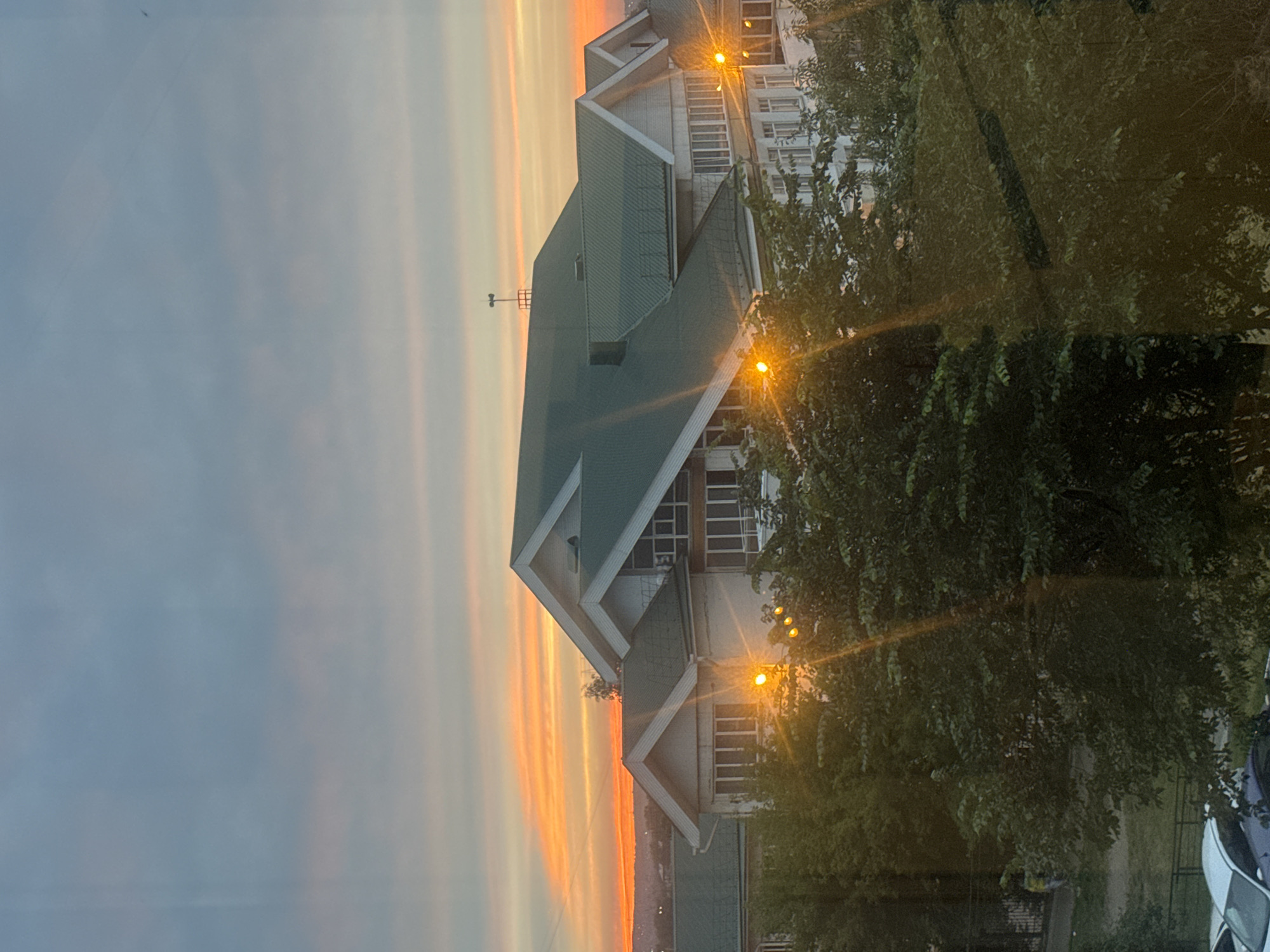 This is a medium shot, eye-level, full-frame photograph of a residential building at dusk in Irkutsk, Russia. The building is multi-story with a distinctive green, pitched roof and white siding. Several windows are visible, some with blinds. In front of the building, trees with dense green foliage are present, with some streetlights reflecting on them. The sky is a gradient of blue and orange, indicating sunset. In the background, there are hints of a distant urban landscape. The overall lighting suggests it is late evening.