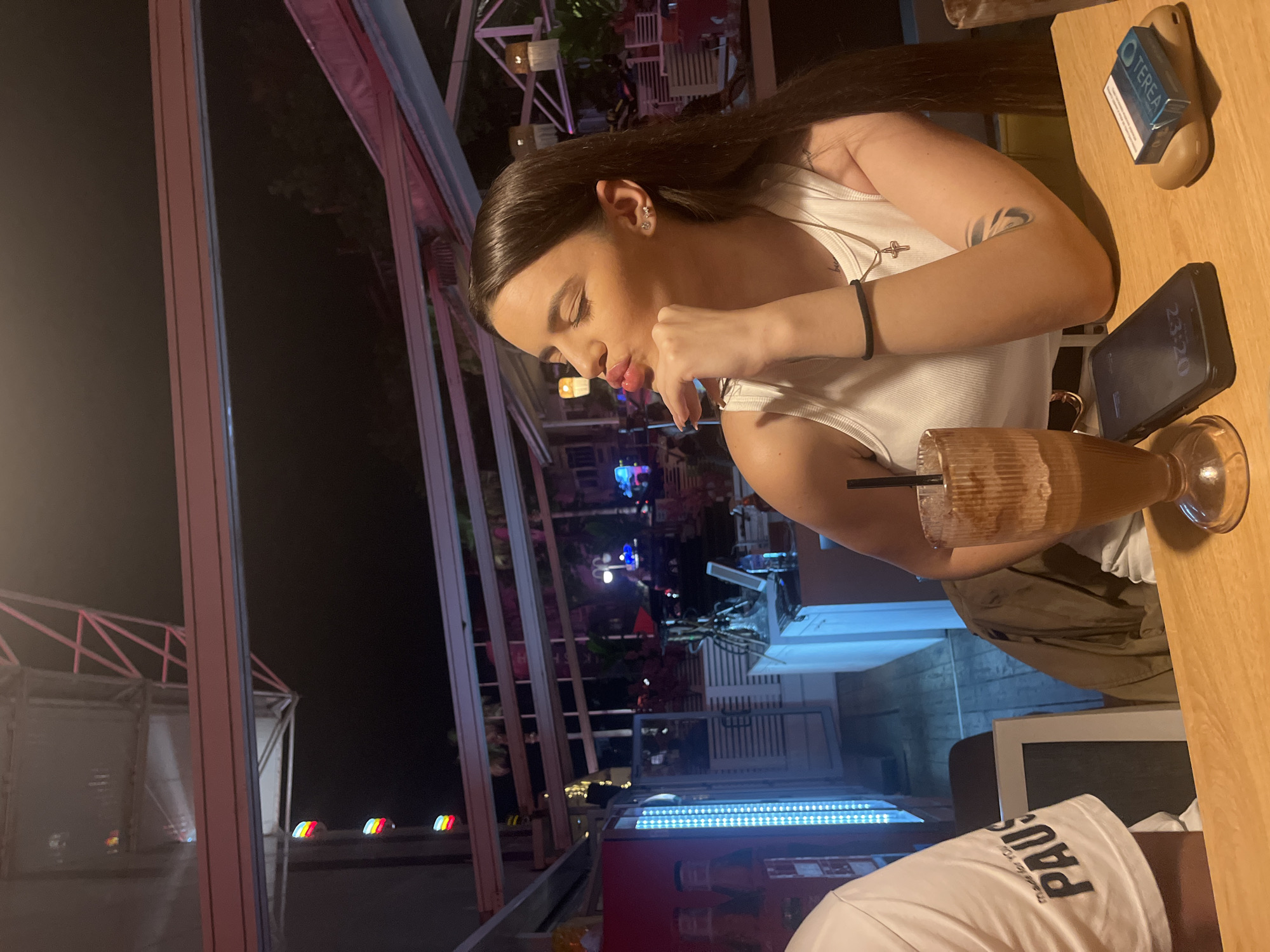 An evening scene depicts a woman and a partial view of another individual seated at a light wooden table within what appears to be an outdoor or semi-outdoor cafe setting at night. The central figure is a young woman with long, dark brown hair, wearing a white ribbed tank top. She has a gold chain with a small cross pendant around her neck and a black elastic band on her left wrist. A tattoo with an abstract, swirling design is visible on her inner left forearm. Her left earlobe features three small stud earrings, one of which is star-shaped. Her head is tilted to the left, her eyes are closed, and her lips are pursed in a pouting gesture, with her right hand, displaying light-colored fingernails, resting under her chin. To her left, the left arm and shoulder of another person are visible, wearing a white t-shirt with black text that includes "PAUS". On the table, a tall glass containing a dark brown, frothy beverage with a black straw is prominently placed in the foreground. A black smartphone, displaying "23:20" on its screen, lies face-up to the right of the woman's drink. Further to the right, a blue pack of "TEREA" brand cigarettes rests on top of a light brown, oval-shaped case or lighter. The background indicates an urban environment at night. Overhead, a structure with white metal beams and what appears to be a translucent or reflective canopy is visible. In the upper left background, and reflected in a surface below it, are distinct vertical striped lights in red, yellow, green, and blue, strongly resembling the Plovdiv Eye Ferris wheel. To the left, a red commercial refrigerator unit features a vertical strip of bright blue LED lights. Faint images resembling stylized figures, possibly Lego-like characters or figures in traditional Middle Eastern attire, are discernible on the refrigerator door. Behind the woman, white slatted panels suggest a bar or partitioned area. A white counter with various bottles, a hookah, and a digital tablet or screen is visible. Further in the distance, ambient lighting from other establishments, trees, and some blue and red flashing lights, possibly from emergency vehicles or entertainment venues, can be seen. The overall lighting scheme is a mix of warm ambient illumination and cooler blue tones from the LED strips. Based on the distinctive colorful vertical lights in the background, which are characteristic of the Plovdiv Eye