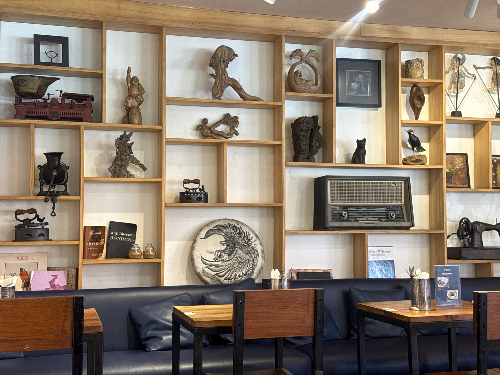 The image depicts the interior of a building, likely a cafe or restaurant, in the Ubud District of Indonesia, as suggested by the menu signage on the tables. The scene is dominated by a large wooden shelving unit filled with various decorative objects, antiques, and artwork. These include sculptures, scales, books, a radio, and framed art. Below the shelves is a dark blue upholstered bench seat with tables and chairs in front. The tables are made of wood with black metal frames, and the chairs have wooden backs and seats. The lighting is bright, and the overall environment appears to be calm and inviting. The scene suggests a space designed to be visually appealing, with a focus on Indonesian art and vintage items.