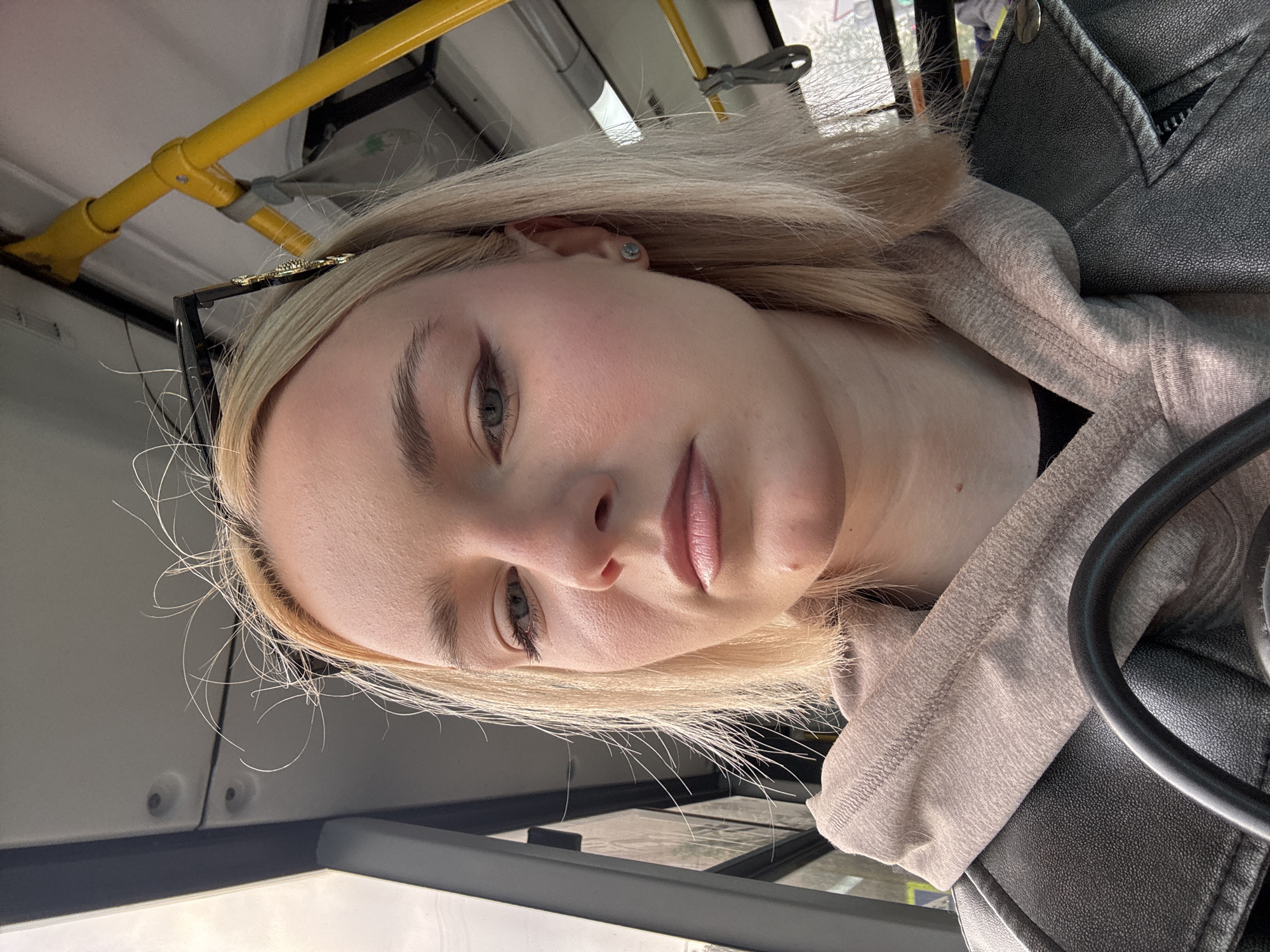 A young woman with blonde hair is taking a selfie in what appears to be a public transport vehicle, likely a bus. She is wearing a beige hoodie layered under a dark leather jacket. Her hair is styled in a bob and some strands are sticking out around her face. She has makeup on, including eyeliner and lipstick. A shiny hair clip is visible in her hair. The interior of the vehicle is visible in the background with yellow handrails and a window. The setting suggests an urban environment.