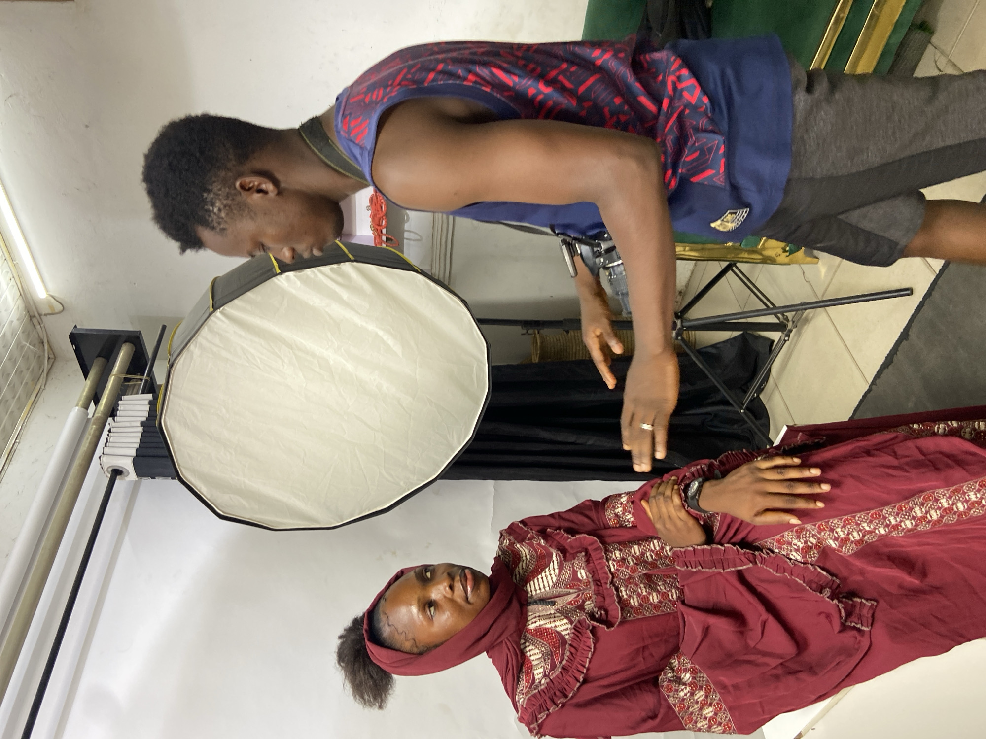 A man is taking a photograph of a woman in a studio setting, possibly in Mararaba, Nigeria. The woman is lying down and wearing a maroon dress with decorative embroidery, as well as a head covering. Her hands are clasped together. The photographer is standing above her, leaning over to capture the image. He is wearing a blue and red patterned shirt and gray shorts, and a camera is visible strapped to his side. A large octagonal softbox is positioned to provide light for the photograph. The background includes a white backdrop, a support system for the backdrop, and various studio equipment. A green chair is partly visible. The floor is tiled.