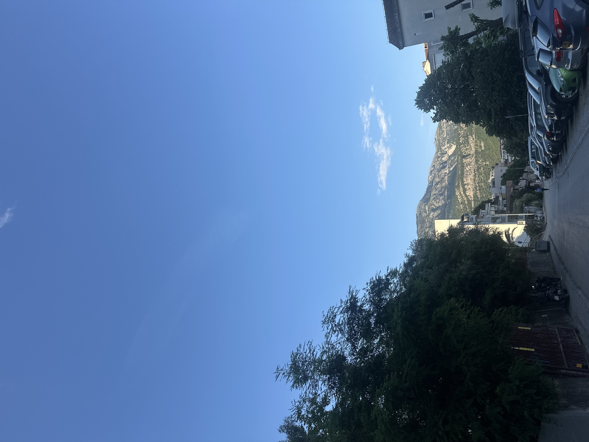 The image shows an outdoor scene in Budva, Montenegro. The sky is mostly clear blue with some clouds. In the distance is a large mountain range. A street is lined with parked cars. Buildings and vegetation are visible along the street. A fence and motorcycles are to the right of the frame.