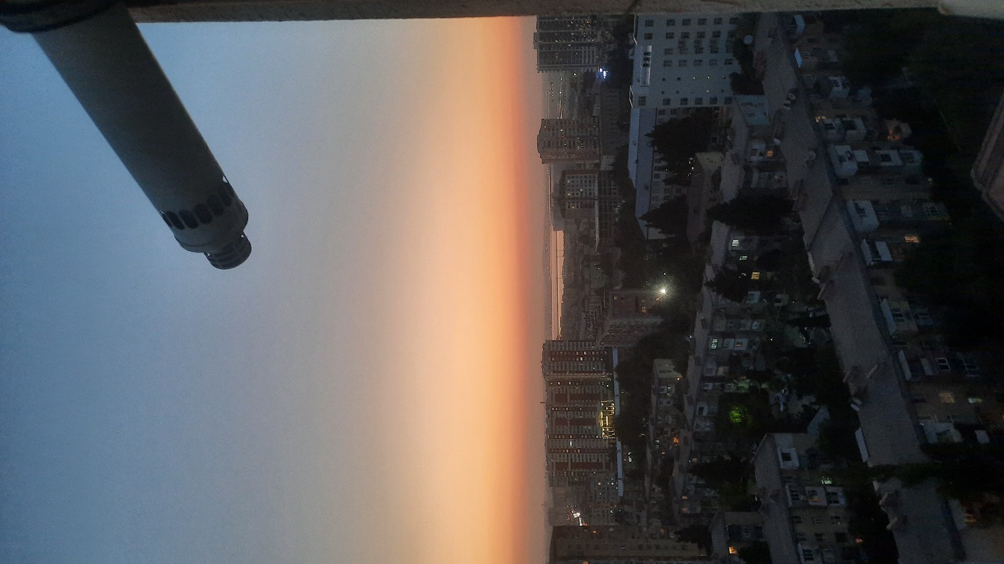 An outdoor, vertical, eye-level shot captures a cityscape at dusk. The sky transitions from a light blue at the top to a vibrant orange-yellow band near the horizon, indicating sunset. In the foreground, a portion of a light-colored pipe extends from the upper right corner, angling downwards into the frame. The pipe has a textured surface with circular perforations near its end. Below the sky, a dense urban landscape unfolds. Numerous apartment buildings of varying heights are visible, some with illuminated windows suggesting occupied residences. The architecture appears to be predominantly multi-story residential blocks. Trees are interspersed throughout the city, some appearing to be deciduous and others evergreen, adding a touch of green to the otherwise muted urban palette. In the distance, a body of water or a large flat expanse is faintly visible beyond the buildings, and further on, a hazy horizon line suggests a vast, open area. The overall atmosphere is one of a transitioning day, with the city lights beginning to twinkle against the fading daylight.
