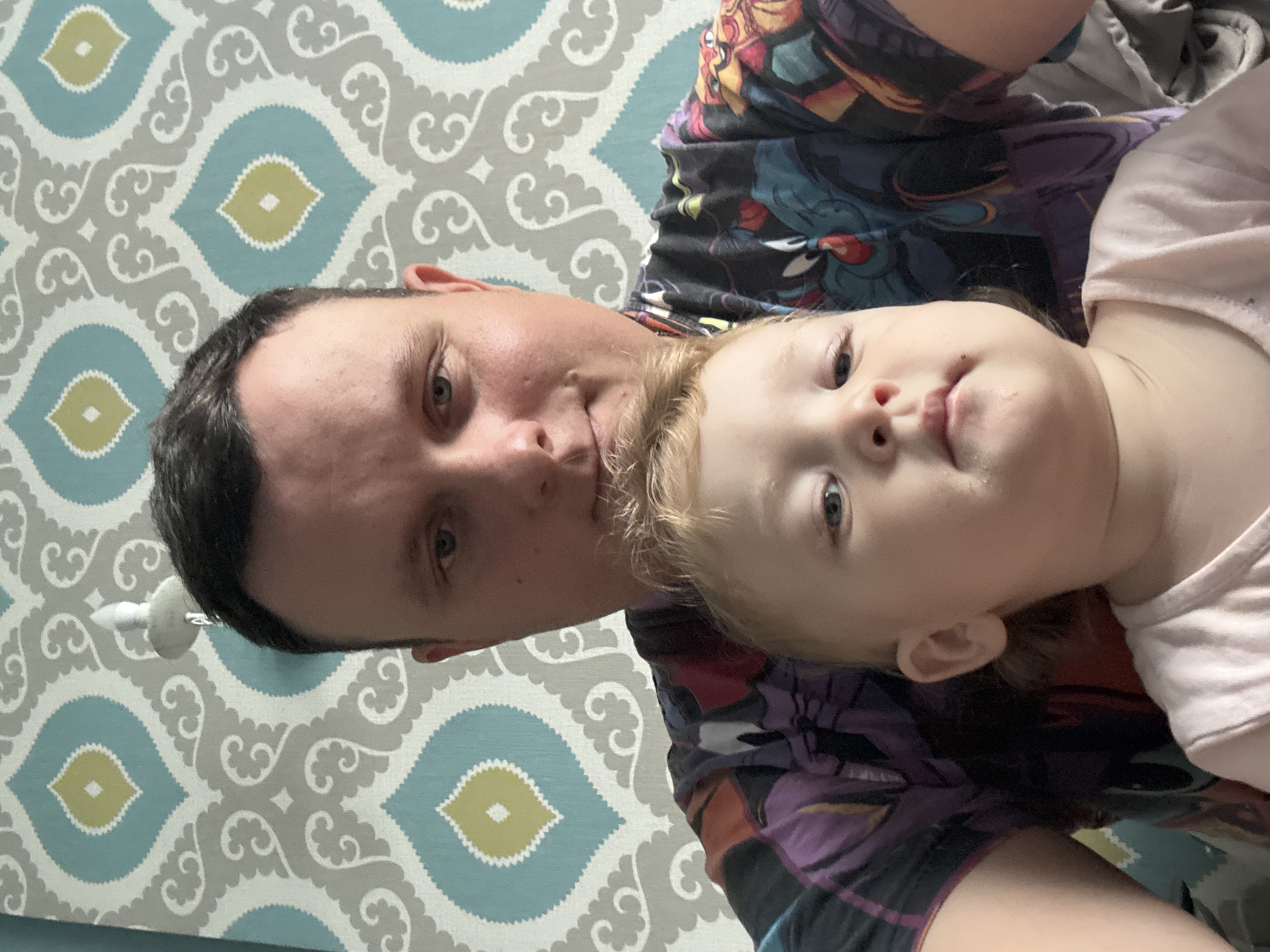The image is a close-up selfie of an adult male and a young child, taken indoors against a backdrop of patterned wallpaper. The adult, positioned behind the child, is looking directly at the camera. The child, in the foreground, is also looking towards the camera. Both are casually dressed. The wallpaper features a repeating pattern of stylized teardrop shapes in teal, grey, and yellow. A light fixture is visible on the wall behind the adult's head. There is no indication of the image being taken in Kondopoga, Russia.