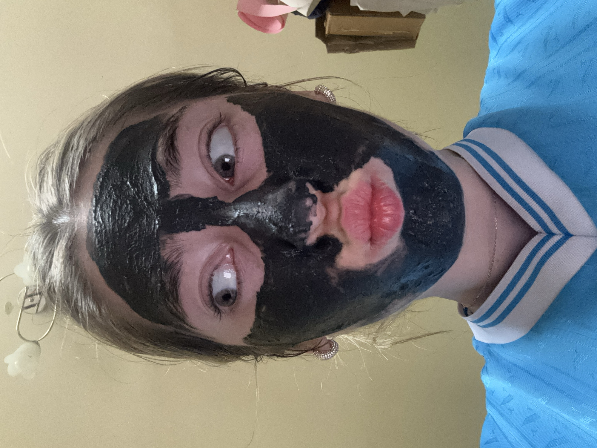 A person with a black face mask is making a kissing face at the camera. The person is wearing a blue shirt with a white collar and has earrings in their ears. The background is a light yellow color, and there is a glimpse of a dresser in the upper portion of the frame. The location in the city of Unknown, Unknown cannot be confirmed.