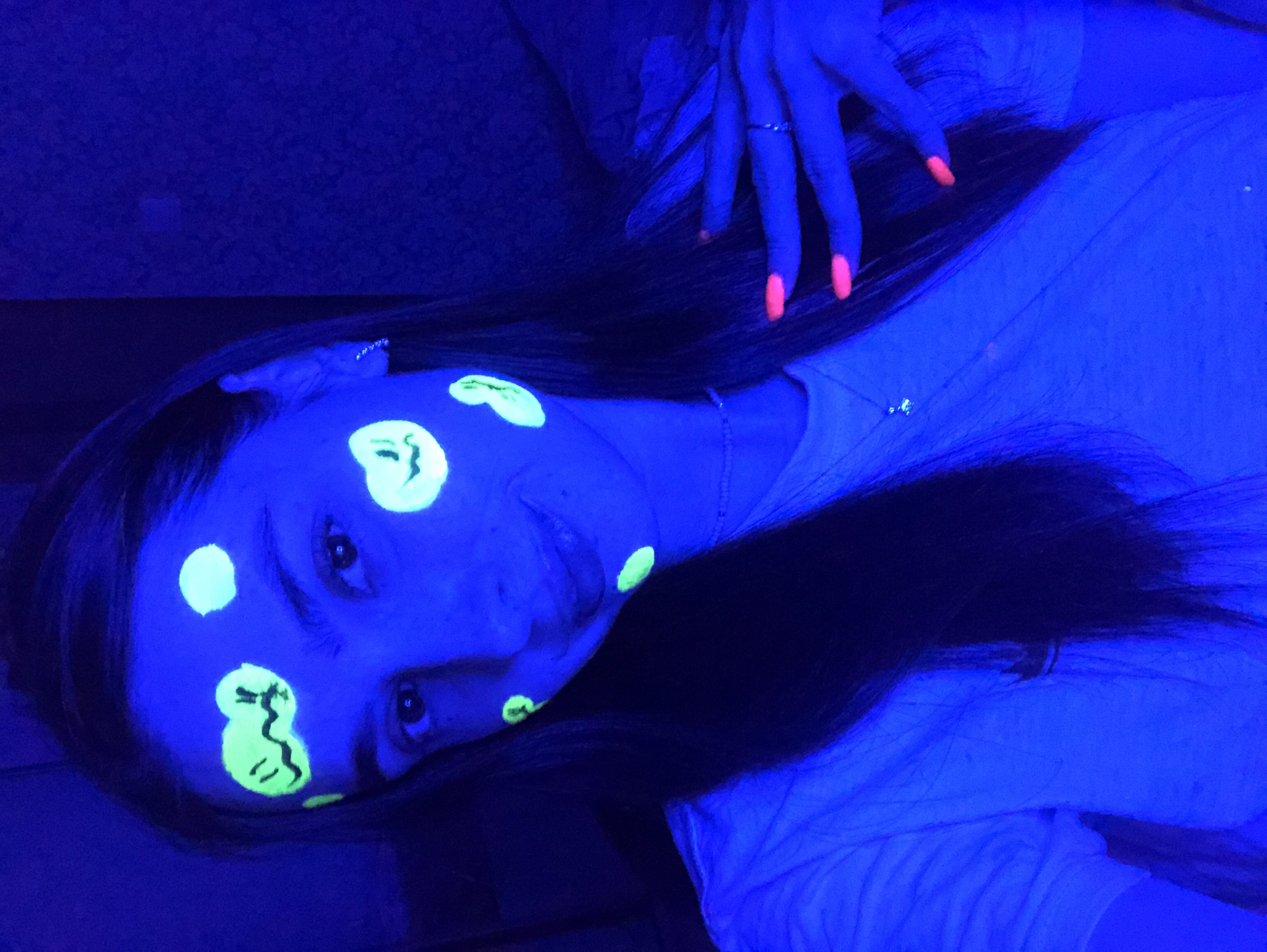 A young woman with long, dark hair poses for a selfie in a dimly lit room, illuminated by a blacklight. Her face is adorned with several small, circular designs in neon green, some resembling smiley faces. Her fingernails are painted a vibrant neon orange. She is wearing a light-colored t-shirt. The background is dark and appears to be a wall or furniture. There are no specific details indicating the location within Noviy, Russia, or any other city.