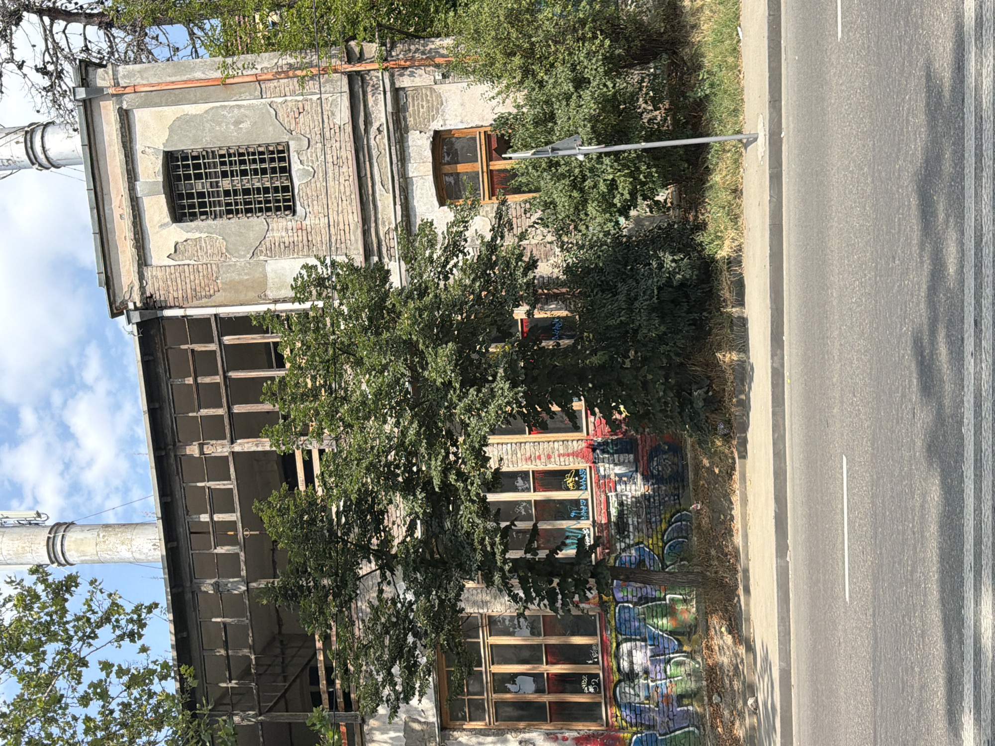 The image shows an abandoned, two-story building on a sunny day in Tbilisi, Georgia. The building has a dilapidated facade with exposed brick and peeling stucco. The windows are either boarded up or broken, and the upper story appears to be a wide, open balcony with wooden beams. Graffiti covers the lower part of the building's exterior walls, and trees and bushes are growing around it. A tall, metallic chimney or pipe rises from the roof. The building is situated next to a paved road with a sidewalk. A street lamp post is visible in front of the building. The sky is blue with scattered clouds.