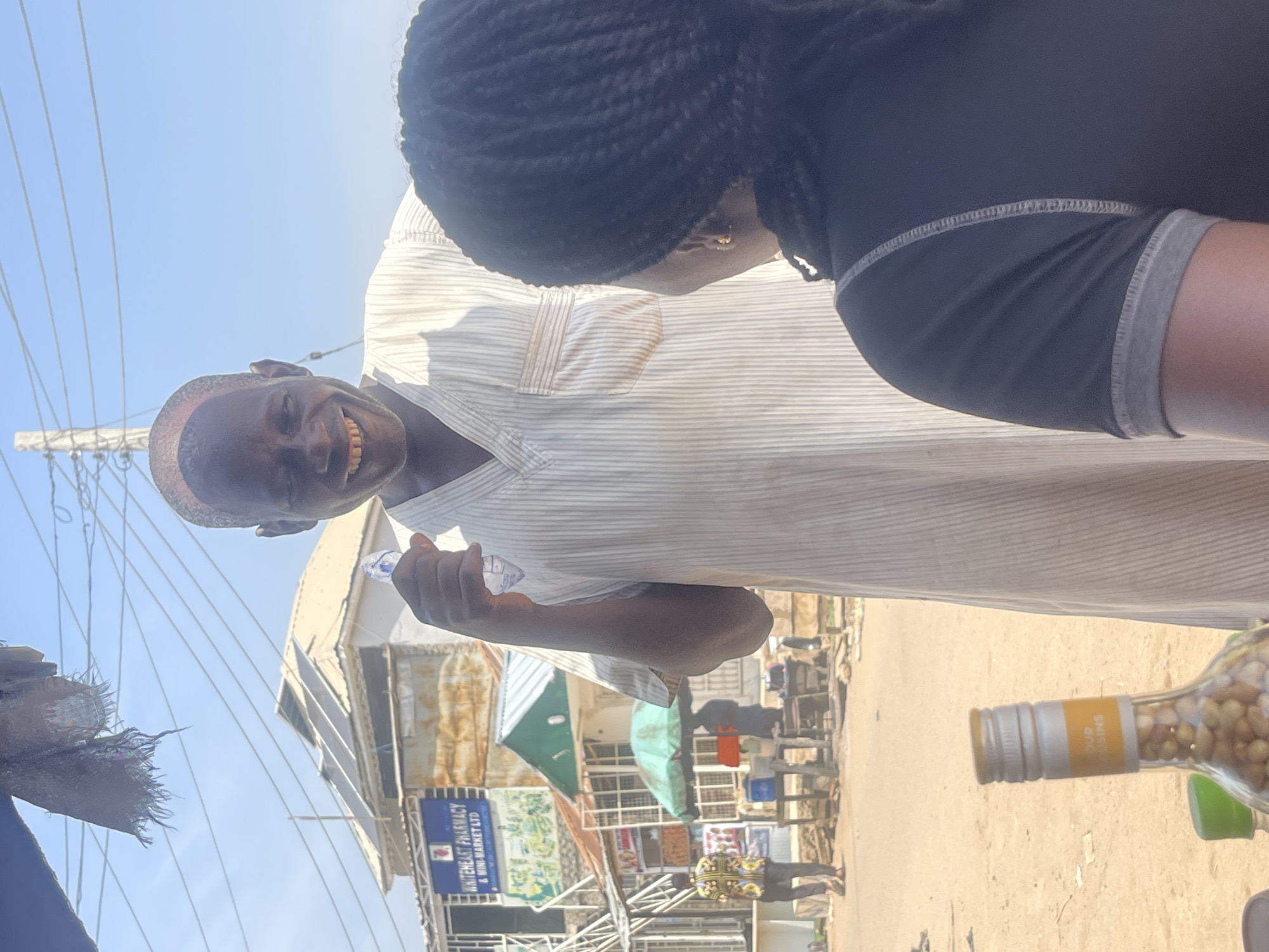 This media file shows a close-up, angled view of two people and a street scene in Abuja, Nigeria. The foreground is dominated by a man's face, captured from a low angle, as he smiles broadly and holds up a plastic bottle. He is wearing a light-colored, striped shirt. Behind him, the back of another person's head is visible, with dark braided hair.
The background reveals a bustling street scene with buildings, market stalls, and people. A prominent sign for "WHITEHEART PHARMACY & MINI-MARKET LTD" is visible, offering a location cue. In the bottom right corner, a glass bottle filled with what appears to be nuts or seeds is partially visible, with a yellow label that has the word "RAISINS" printed on it. The overall impression is a candid snapshot taken outdoors during daylight, possibly in a market area. The bright sky suggests a sunny day. The man's expression conveys happiness or amusement, and the scene has a lively, everyday feel typical of a busy urban environment.