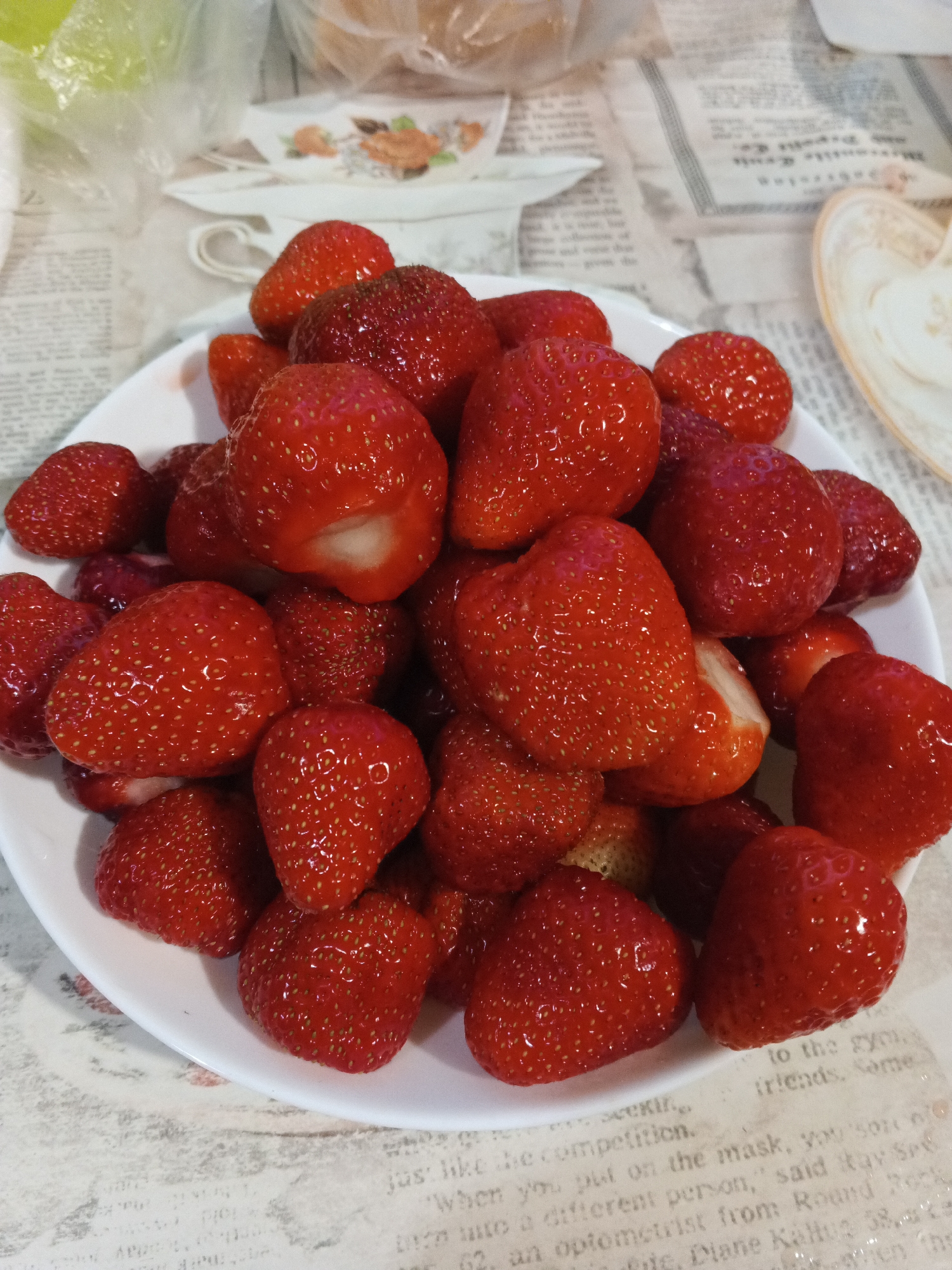 The image shows a white plate filled with ripe strawberries, a close-up shot with a shallow depth of field. The strawberries are a vibrant red, with visible seeds and some showing the white interior where they have been cut. They are piled high on the plate, filling most of the frame. In the background, a surface that appears to be covered in newspapers or printed paper is visible. The text on the paper is partially obscured but suggests a casual setting, possibly a kitchen or dining area. The lighting is soft and even, indicating it could be daytime or well-lit indoor lighting. There are no people or animals visible in the image. The focus is entirely on the fresh fruit. The overall impression is one of simple abundance and freshness.