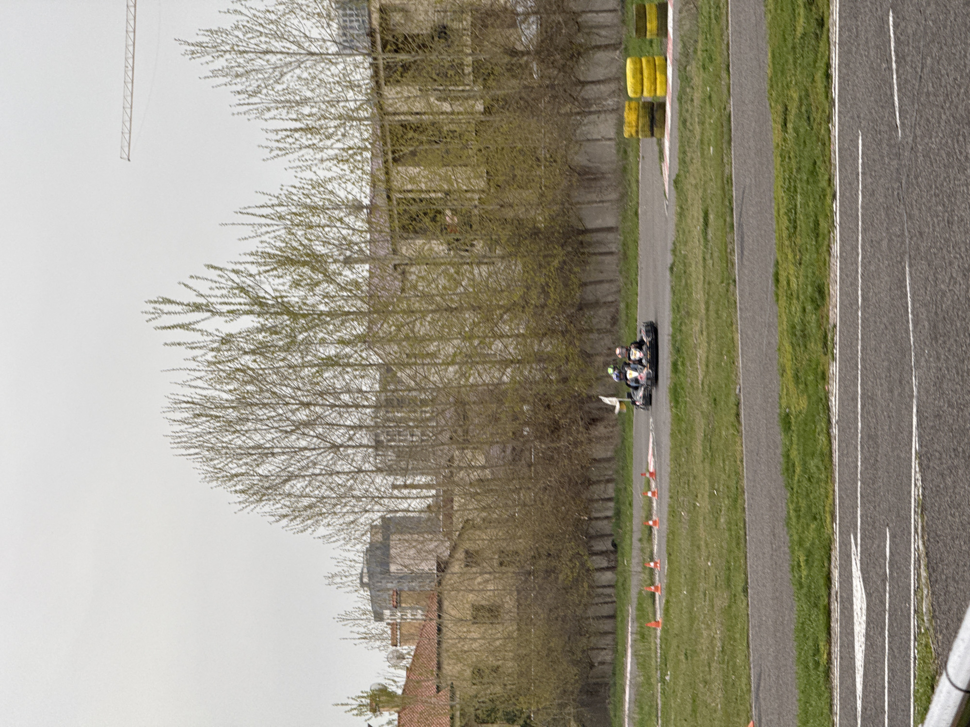 The image depicts a go-kart race taking place on a track. Two go-karts are visible, with drivers wearing helmets. The track has grass bordering it, marked by orange cones along its edge. There are yellow and black barriers on the track. Beyond the track are trees and some buildings, and a crane can be seen in the upper left of the frame. The photograph suggests an overcast day. The location is in Yerevan, Armenia.