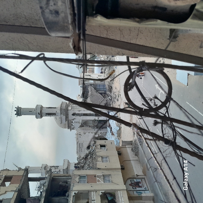 The image depicts a devastated urban streetscape in Khan Yunis, Palestinian Territory, under an overcast sky. The viewpoint appears to be from an elevated position, possibly through a damaged railing or structure composed of tangled metal wires and rebar in the immediate foreground, with a partial view of a building facade on the right.
To the left, a multi-story building exhibits extensive structural damage, particularly on its upper floors where large sections are missing, revealing exposed rebar and interior debris. Balconies on lower, less severely damaged floors show laundry hanging. The ground floor of this building features a sign with a red triangle, circle, and square, and the text "DELTA" and "PLAYSTATION GAMES."
A paved road runs through the midground, marked with a white lane and a white arrow indicating a turn to the left. Debris and rubble line both sides of the road. Two small, indistinct figures are visible walking on the road in the mid-right, moving away from the viewer; one appears to be carrying an object.
In the background, a prominent mosque, identifiable as the Great Mosque of Khan Yunis, stands with significant damage. Its main dome and tall minaret show multiple impact holes, shattered sections, and structural compromise. Adjacent and behind the mosque, numerous other buildings are visible in varying states of destruction, reduced to rubble or damaged shells. The overall environment suggests widespread devastation.