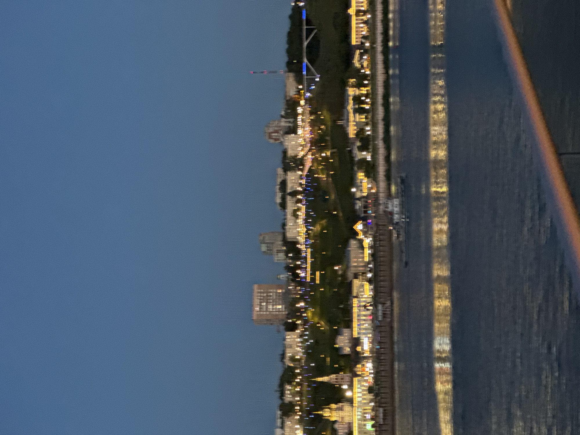 This is a vertical, nighttime photograph of a city skyline along a body of water, likely a river. The image is shot from a high vantage point, possibly from a bridge or tall building. The city is illuminated by numerous lights from buildings, streets, and possibly a Ferris wheel or other amusement park attraction visible in the distance. The lights are reflected in the dark water, creating a shimmering effect. The sky is a deep blue, indicating dusk or early night. The overall mood of the image is serene and picturesque, capturing the beauty of a city at night. Some of the buildings appear to be residential, while others might be commercial or public structures. The presence of a waterfront suggests a lively urban area with potential for recreational activities. The scene is likely in Nizhny Novgorod, Russia, given the context. There is no visible text in the image.