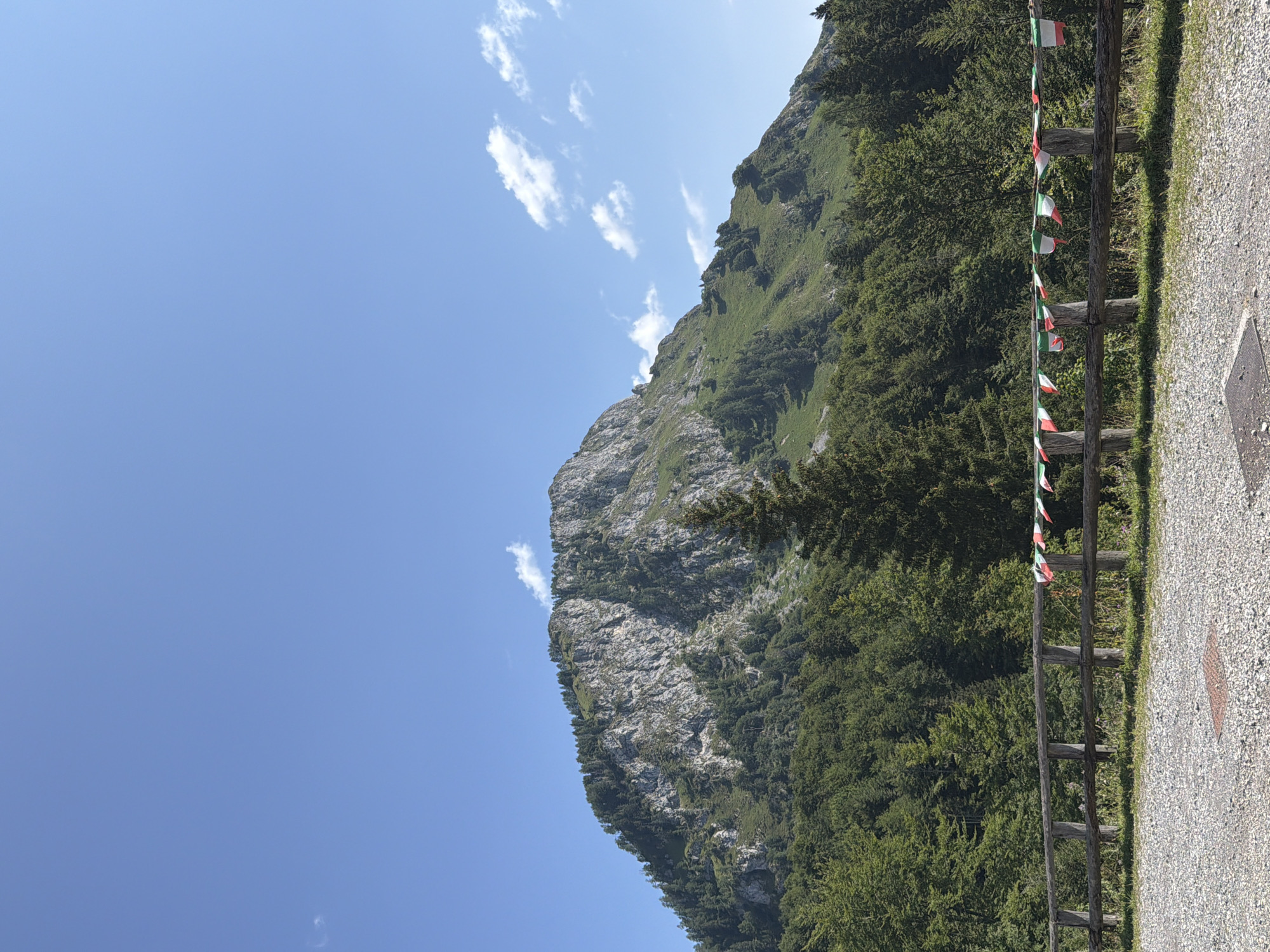 The image depicts a panoramic view of a mountainous landscape in Ampezzo/Dimpeç, Italy. In the foreground, a rustic wooden fence runs horizontally across the frame, adorned with a string of small Italian flags. Beyond the fence, a dense forest of evergreen and deciduous trees covers the lower slopes of a large, rocky mountain. The mountain's summit is rugged and appears to be partially covered in scree. The sky above is a clear, vibrant blue, with a few wispy white clouds scattered across it. The ground in the immediate foreground consists of gravel. The scene suggests a clear, sunny day in a mountainous region.