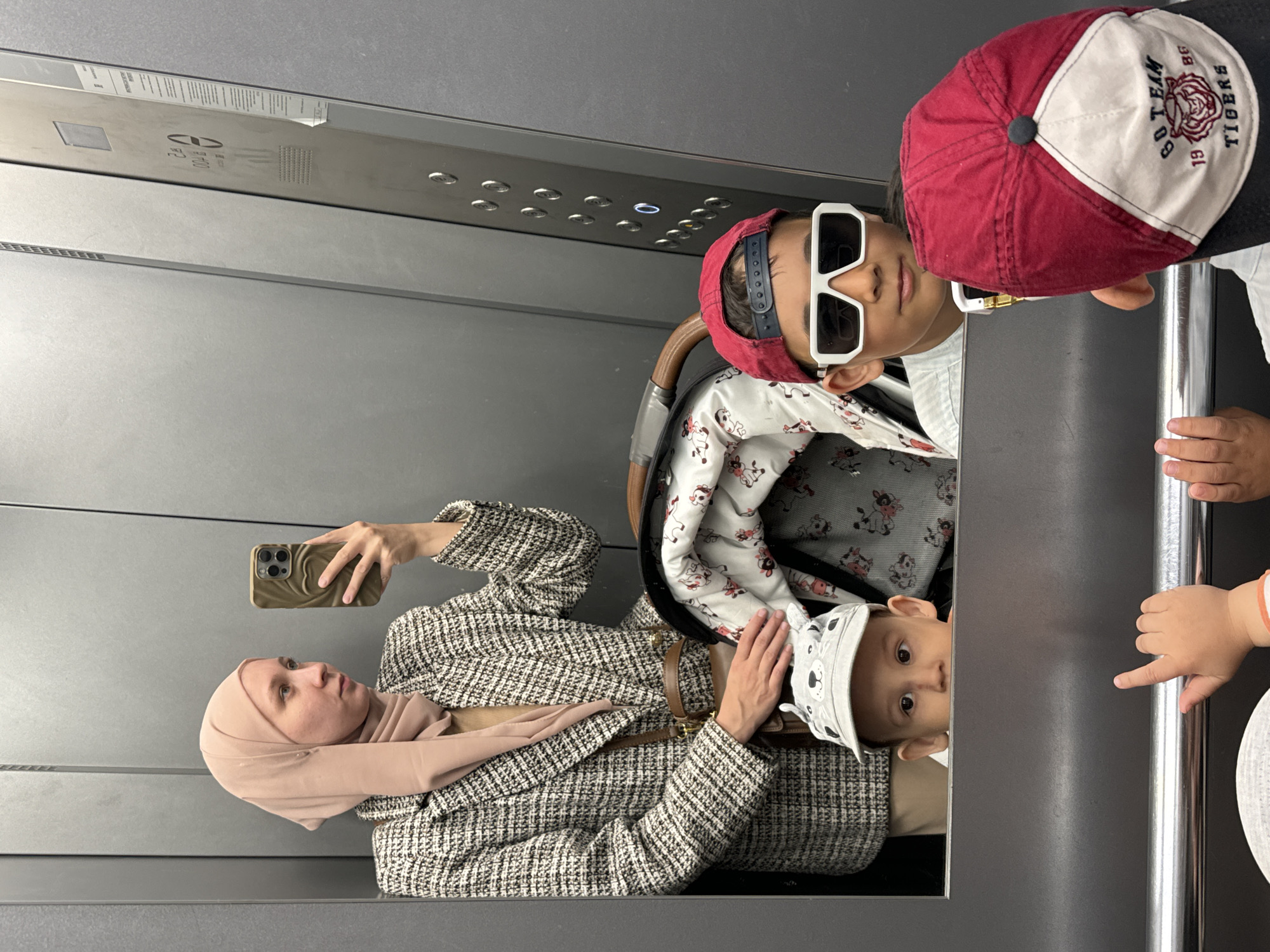 The image shows a woman wearing a tan hijab and a brown and white tweed jacket, taking a selfie with a smartphone inside an elevator. With her is a baby in a stroller, and two other children. One child wears white sunglasses and a red baseball cap; the other wears a red and white baseball cap. The elevator has grey walls and buttons. The location is unspecified, and there is no mention of Chaikovsky, Russia, in the image.